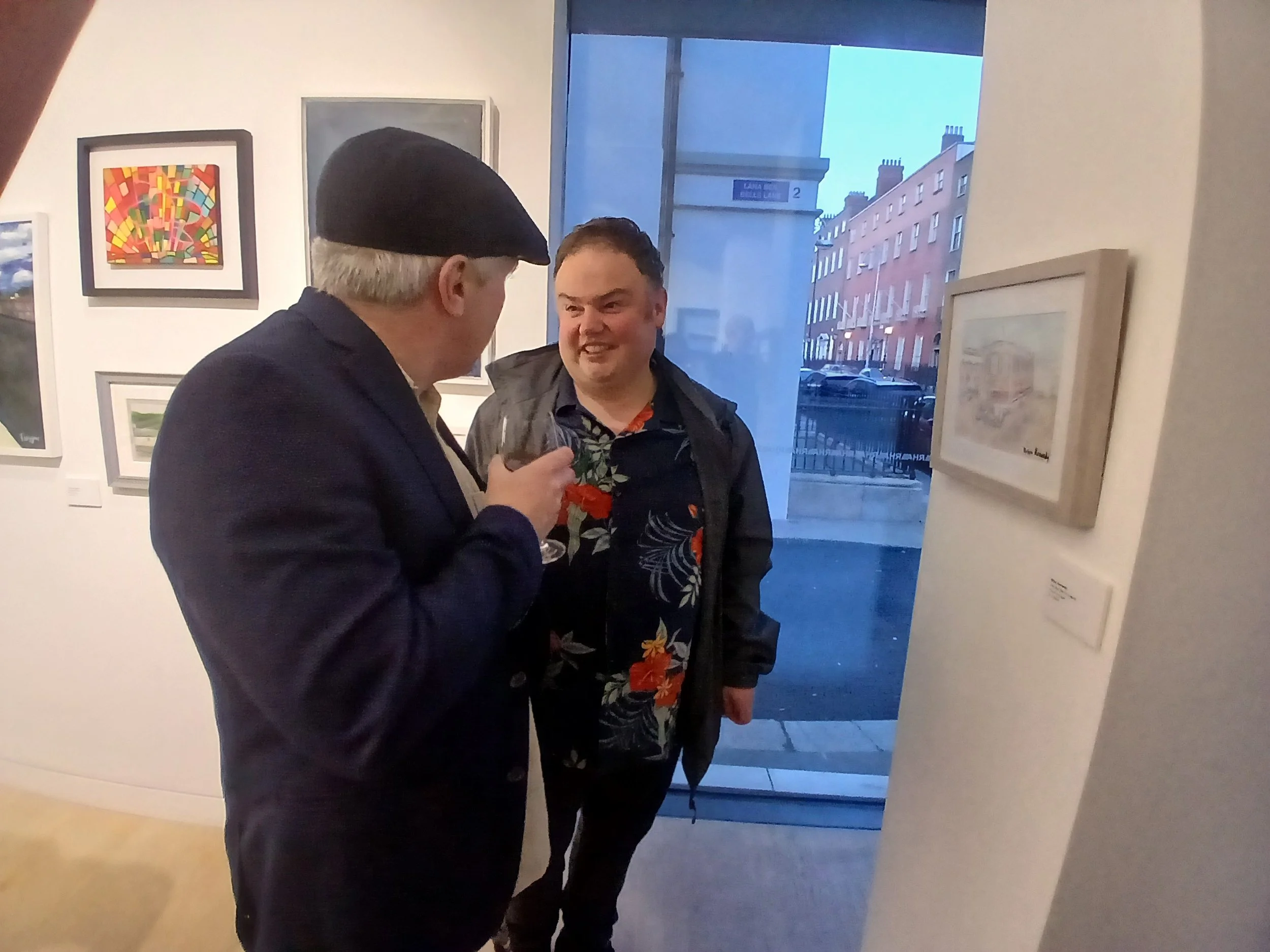 Two individuals converse in an art gallery, with one wearing a dark flat cap and suit jacket, holding a wine glass, and the other in a floral shirt and jacket. Various artworks hang on the white walls behind them, and a window offers a view of a stre