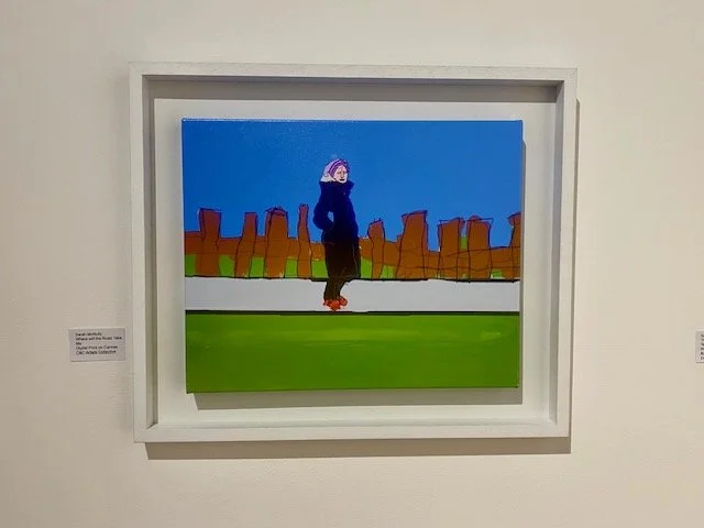 A framed colorful digital print on canvas depicts a solitary figure in a dark coat and purple hat standing on a gray path against a bright blue sky and a fence made of rough brown posts.