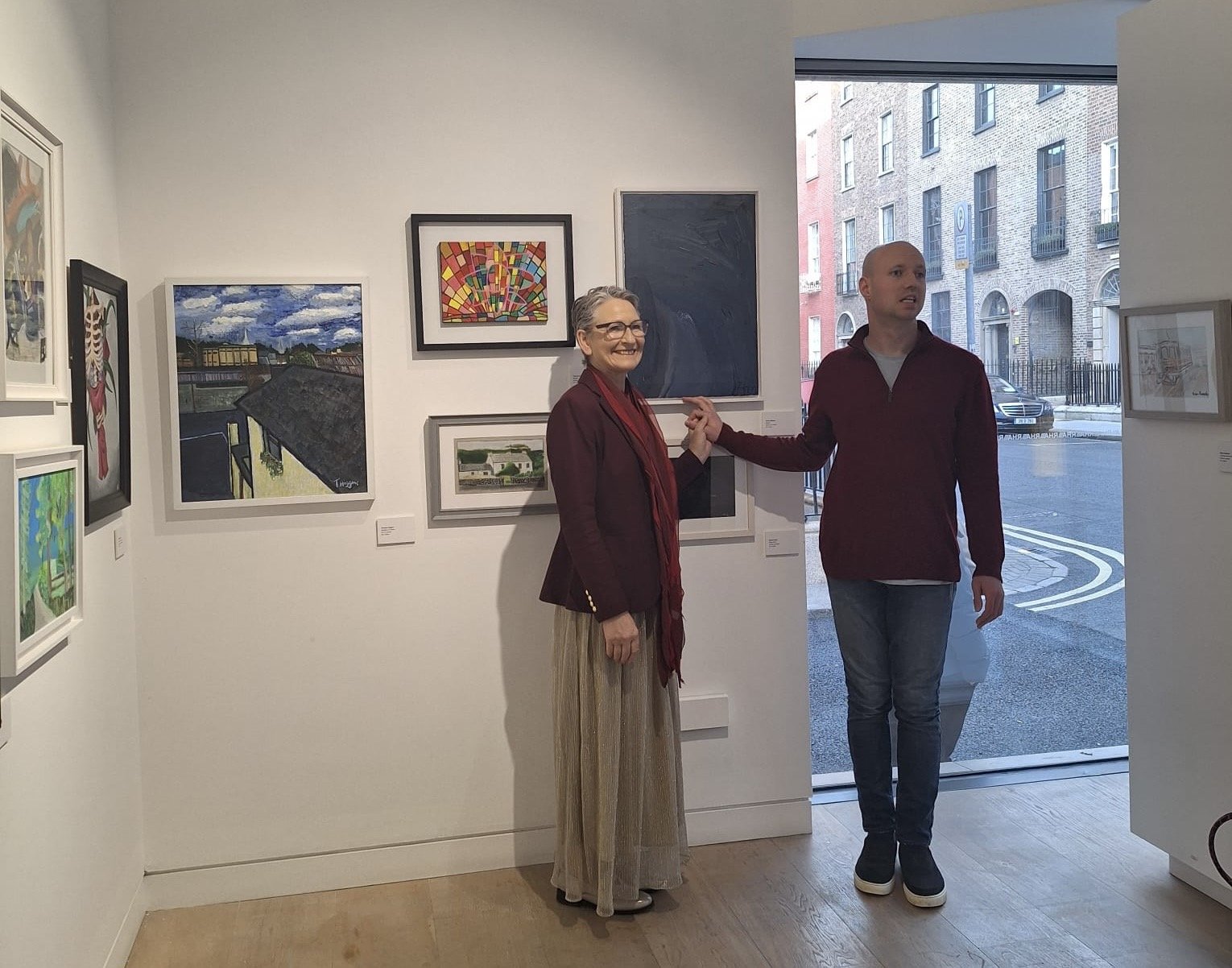 Two individuals stand in an art gallery with paintings displayed on the white walls and a street scene visible through a large window.