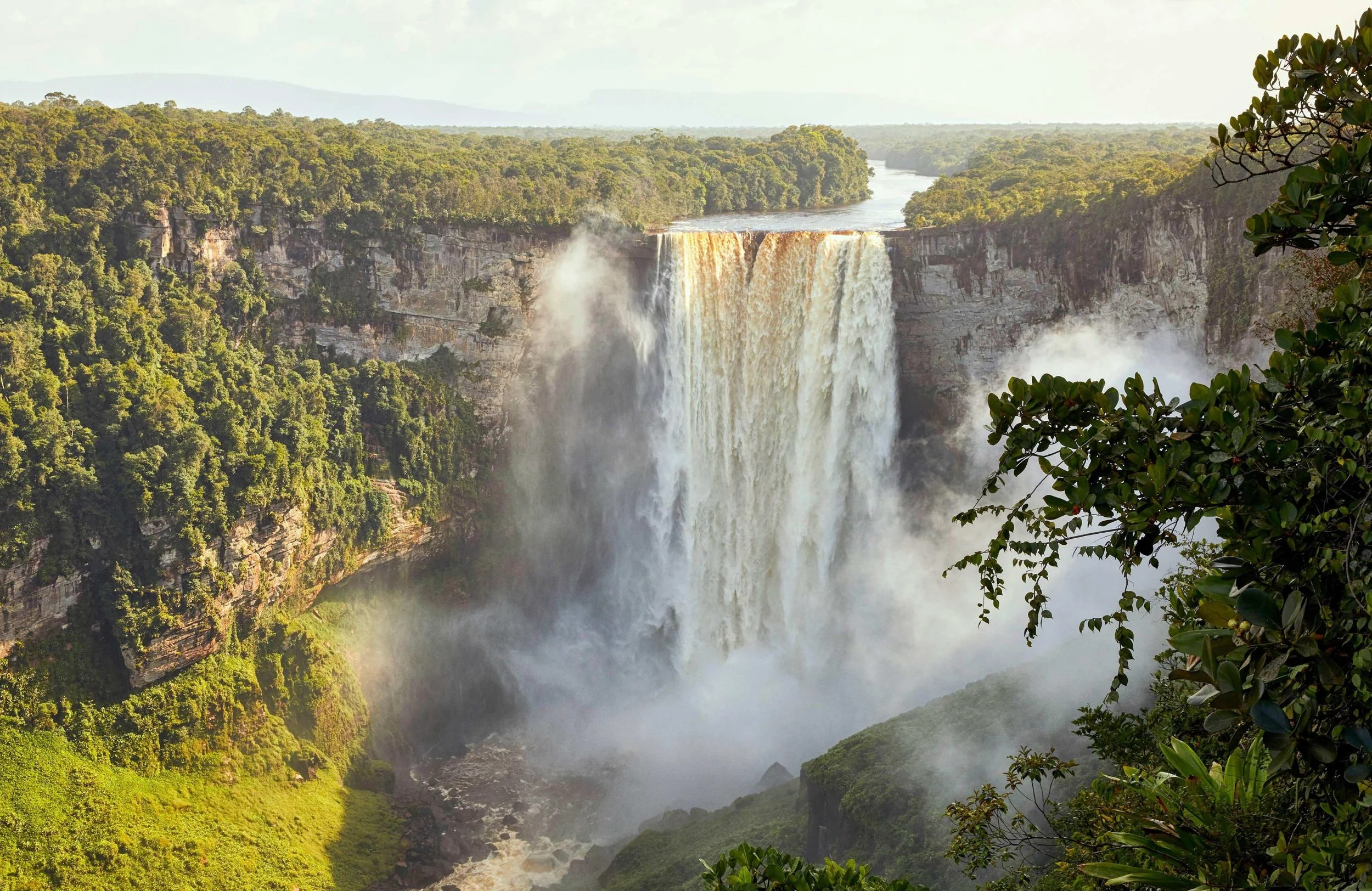 A large waterfall cascading into a misty river below, surrounded by lush, green forest and trees on both sides.