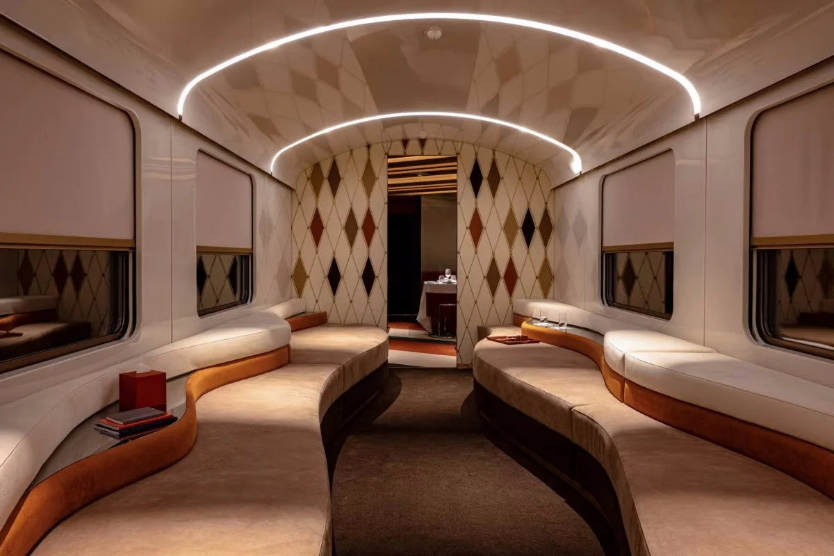 Luxurious train carriage with curved beige and brown seating along the walls, modern lighting, and a patterned wall at the far end.