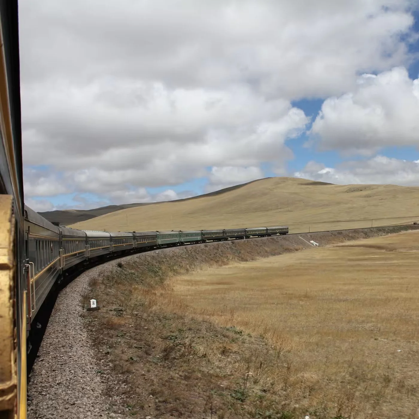 A train traveling through a rural landscape with yellowish grass, rolling hills, and a partly cloudy sky.