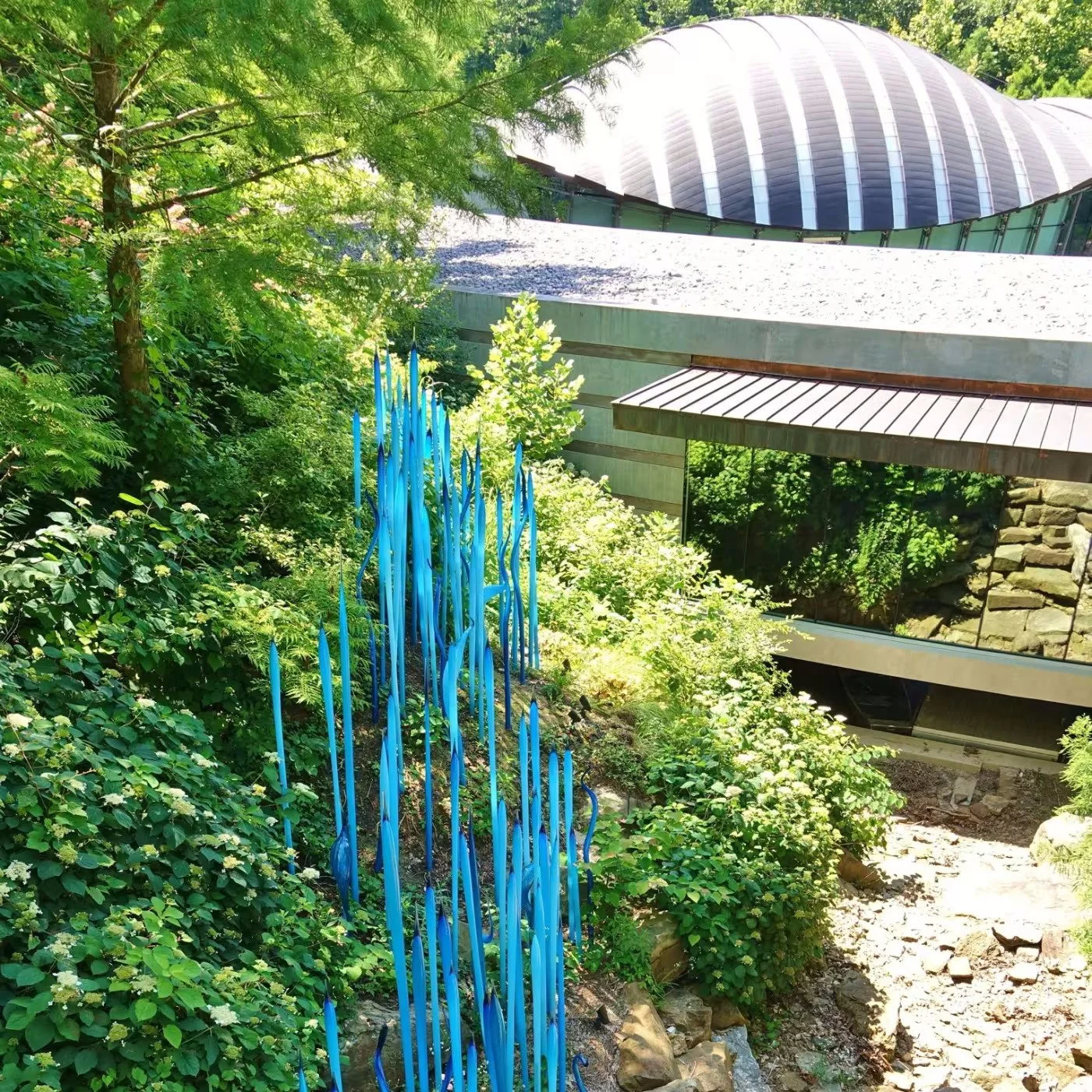 Green trees and bushes surround a modern building with a curved roof, glass windows, and metal siding. A blue glass art sculpture resembling flame tips is on a hillside among the plants.