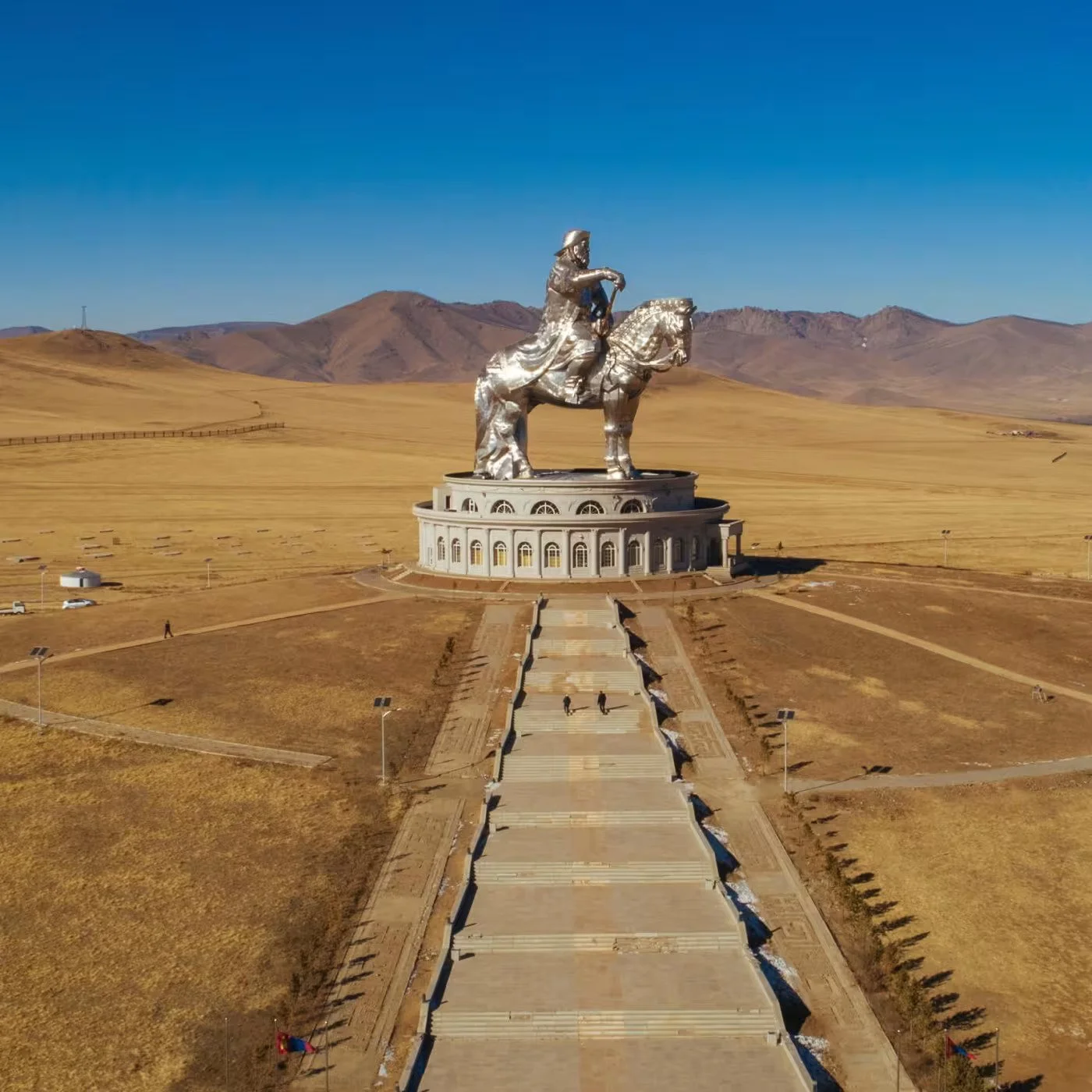 An aerial view of a large metallic statue of a man on a horse in a vast open landscape with mountains in the background, accessible via wide stairs and surrounded by a park area.