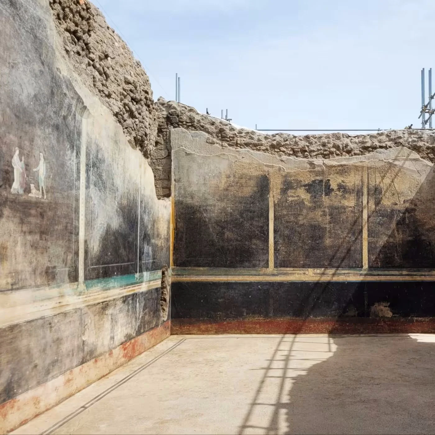 Ancient frescoes on the walls of an archaeological site, with exposed stonework above. The frescoes depict figures and scenes, some partially preserved, on a black background.