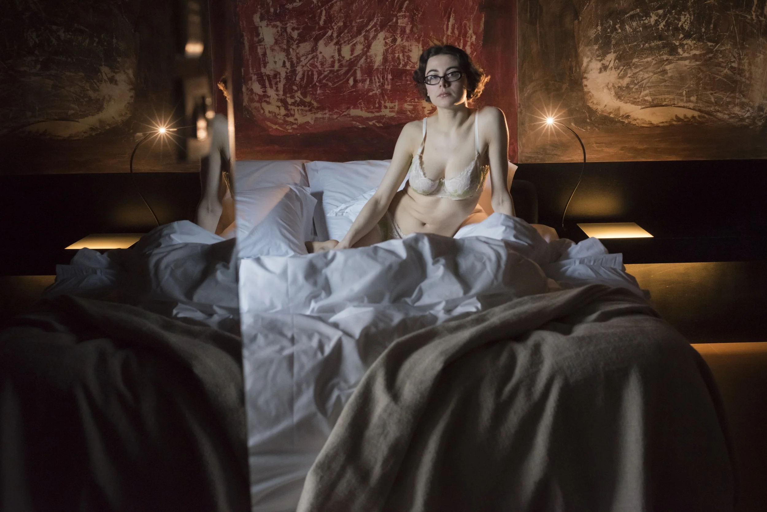 A woman in lingerie sitting on a bed in a dimly lit room with abstract wall art and bedside lamps.