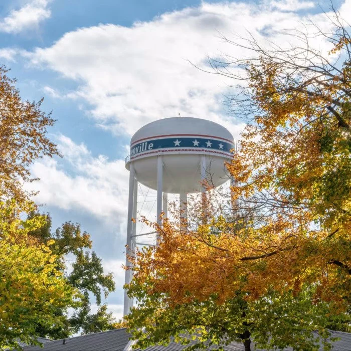 A water tower with patriotic blue, white, and red stripes and stars, seen through trees with autumn leaves, under a partly cloudy sky.