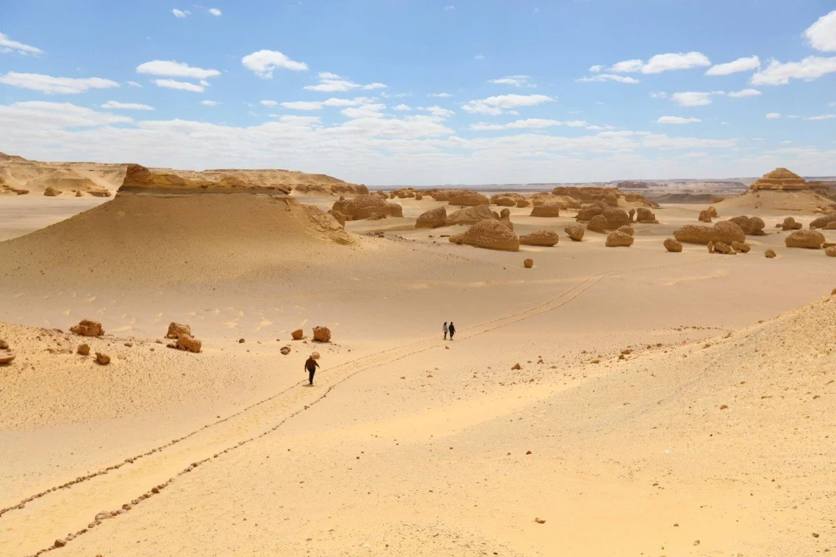 Three people walking through a vast desert landscape with large rock formations under a partly cloudy sky.