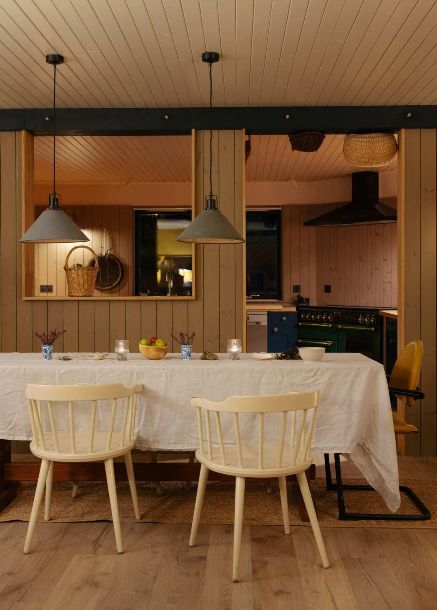 Dinner table set with a white tablecloth, small potted plants, candles, fruit bowl, and dishes in a rustic kitchen with wood-paneled walls and ceiling, pendant lights, and interior windows.