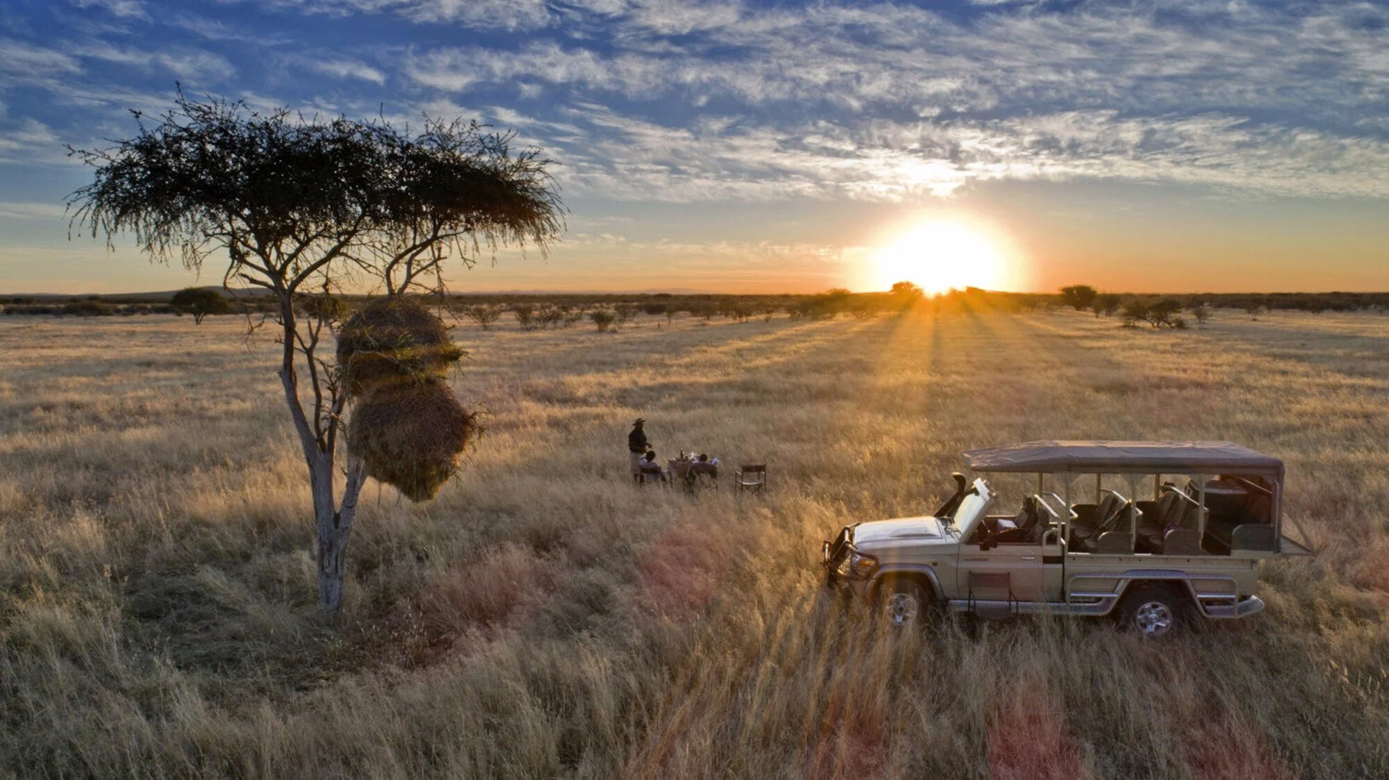 A safari scene at sunset with a rugged vehicle parked on tall grass. In the distance, a person stands near a small table and chairs, toward the horizon where the sun is setting. A large tree with sparse leaves is in the foreground, and the sky is partly cloudy with rays of sunlight shining through.