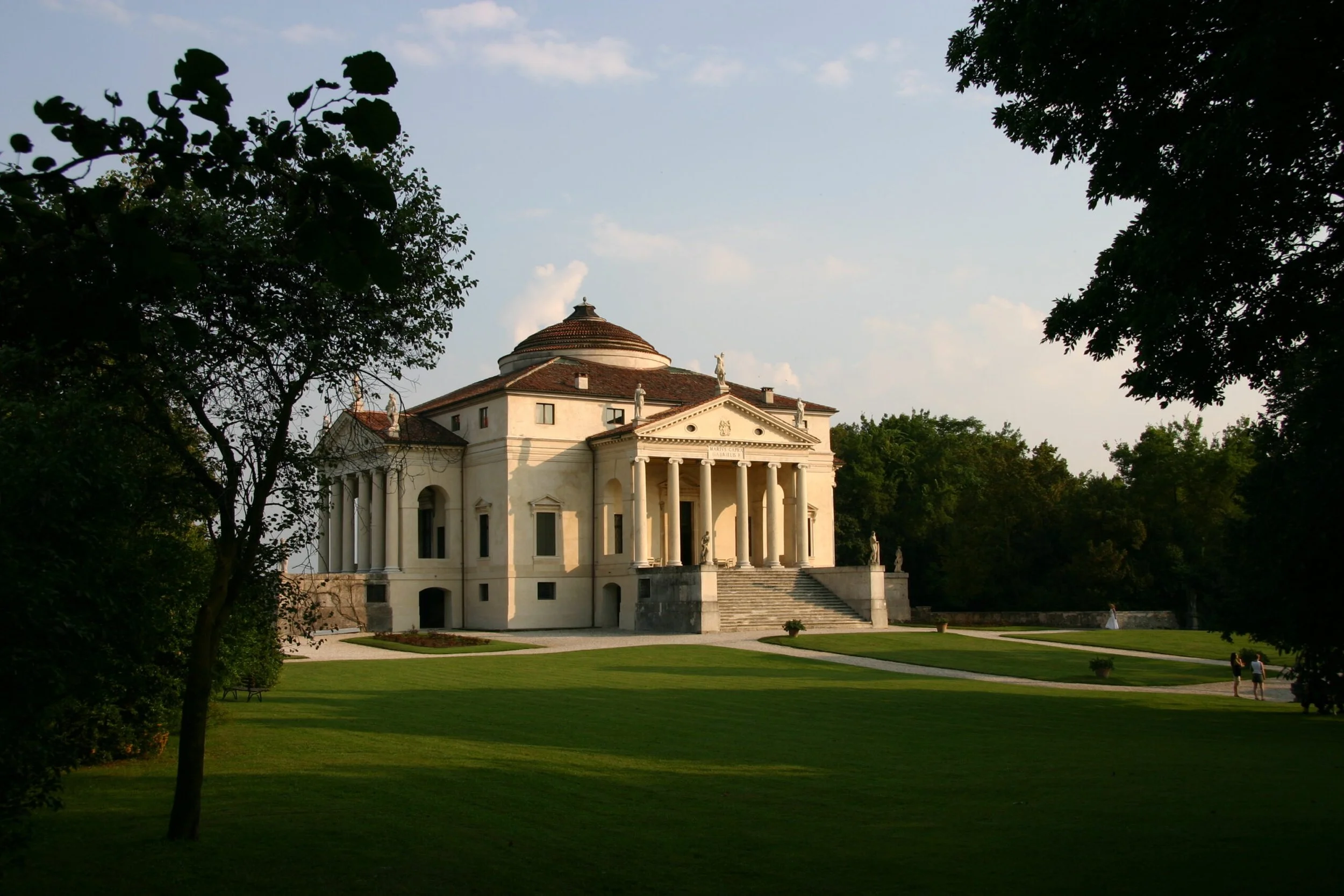 A grand neoclassical building with a dome, columns, and statues on the roof, situated on a well-manicured lawn with trees on either side and a few people walking nearby.