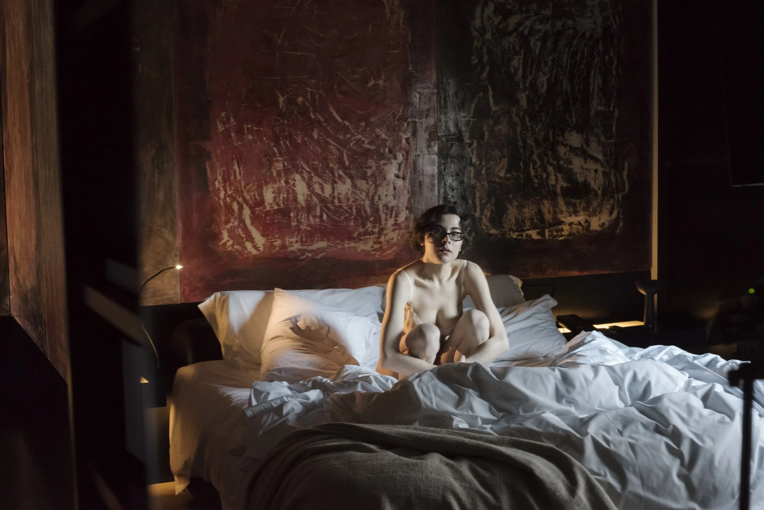 A woman with glasses sitting on a bed in a dimly lit bedroom, with a dark, abstract painting on the wall behind her, white sheets and pillows on the bed, and a warm, intimate atmosphere.