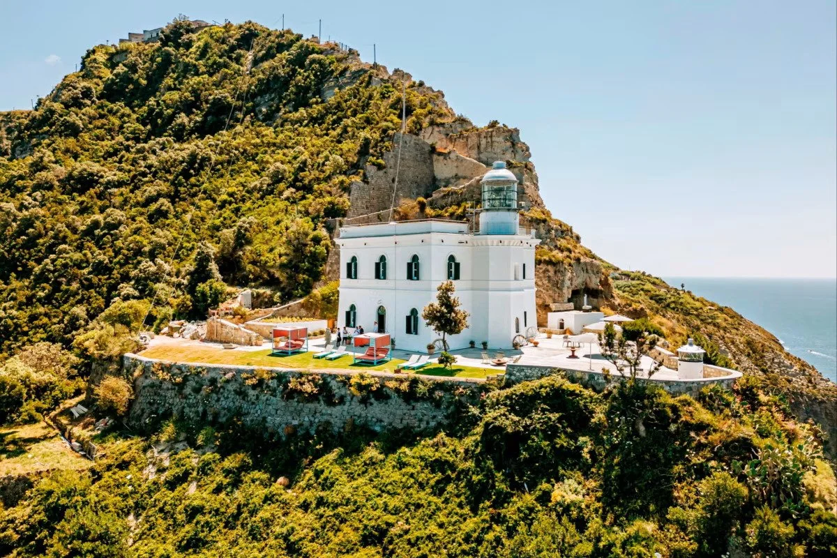 A white lighthouse with a small building at its base sits on a rocky hillside surrounded by trees, overlooking the ocean, with a clear blue sky overhead.