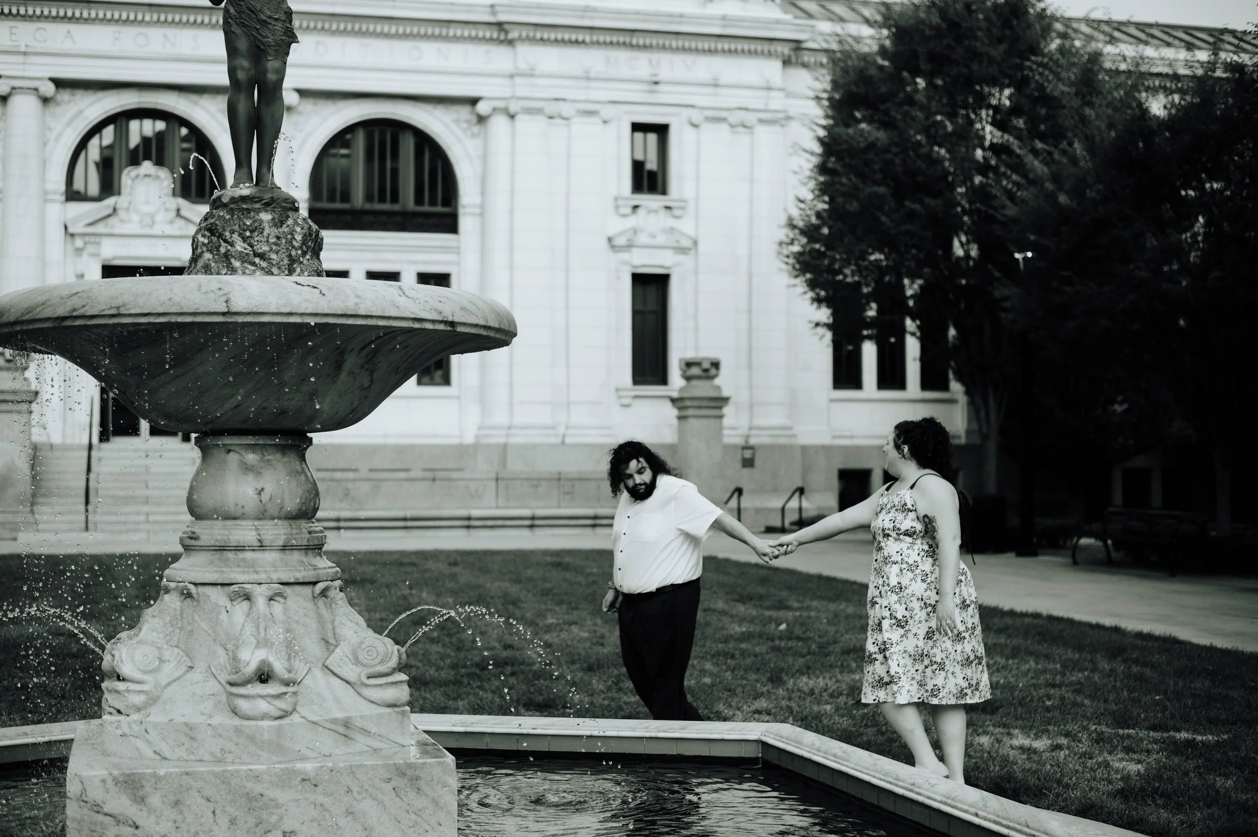 A black and white photo of a man and woman holding hands near a fountain outside a historic building, with trees and benches in the background.