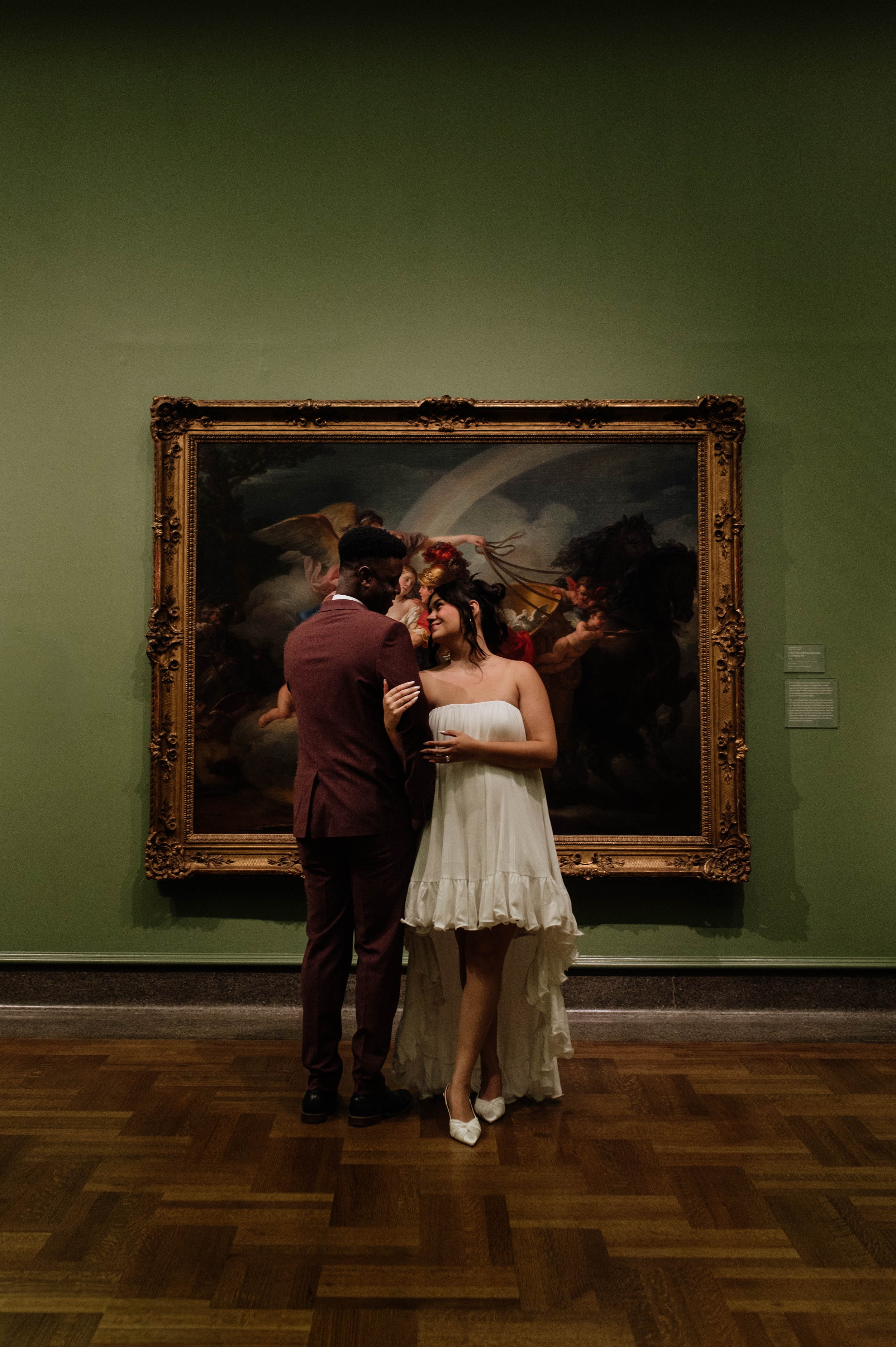 A couple dressed in formal attire standing close and smiling at each other in front of a large, ornate framed painting in an art gallery.