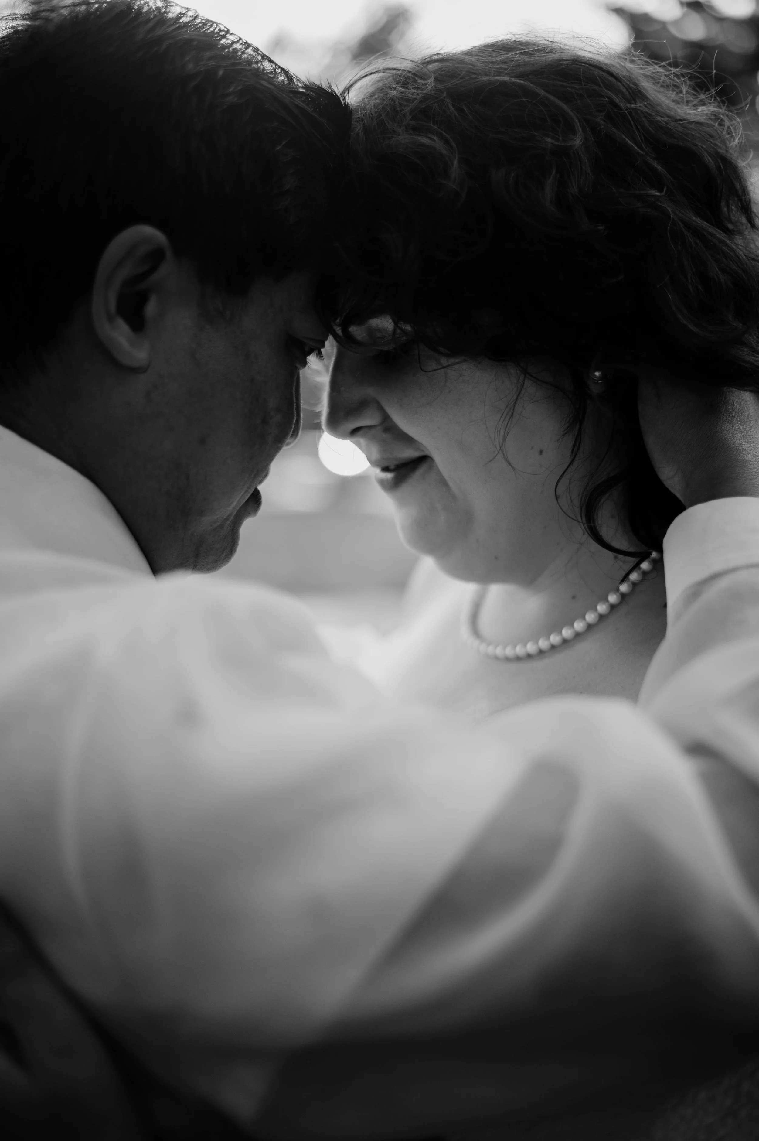 A black-and-white photo of a man and woman gently touching foreheads, eyes closed, sharing an intimate moment. The woman is wearing a pearl necklace and earrings, and the man has short dark hair.