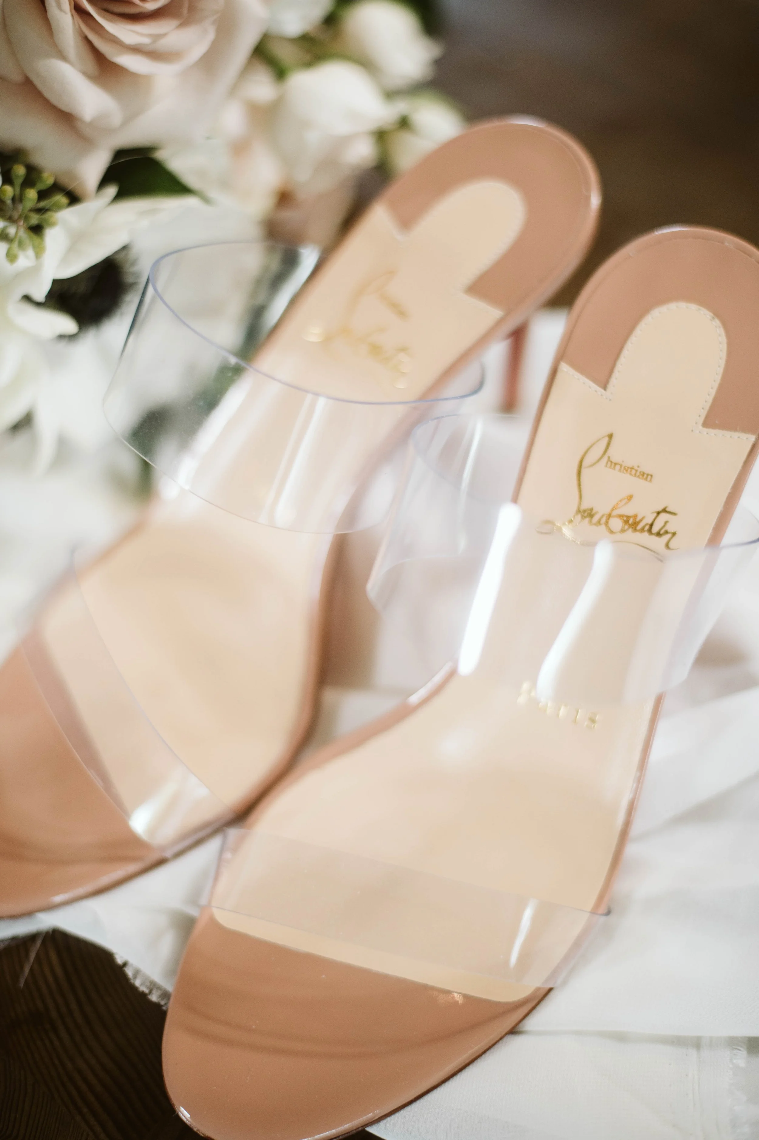 A pair of Christian Louboutin high-heeled shoes with clear straps and blush pink soles, adorned with a floral arrangement.