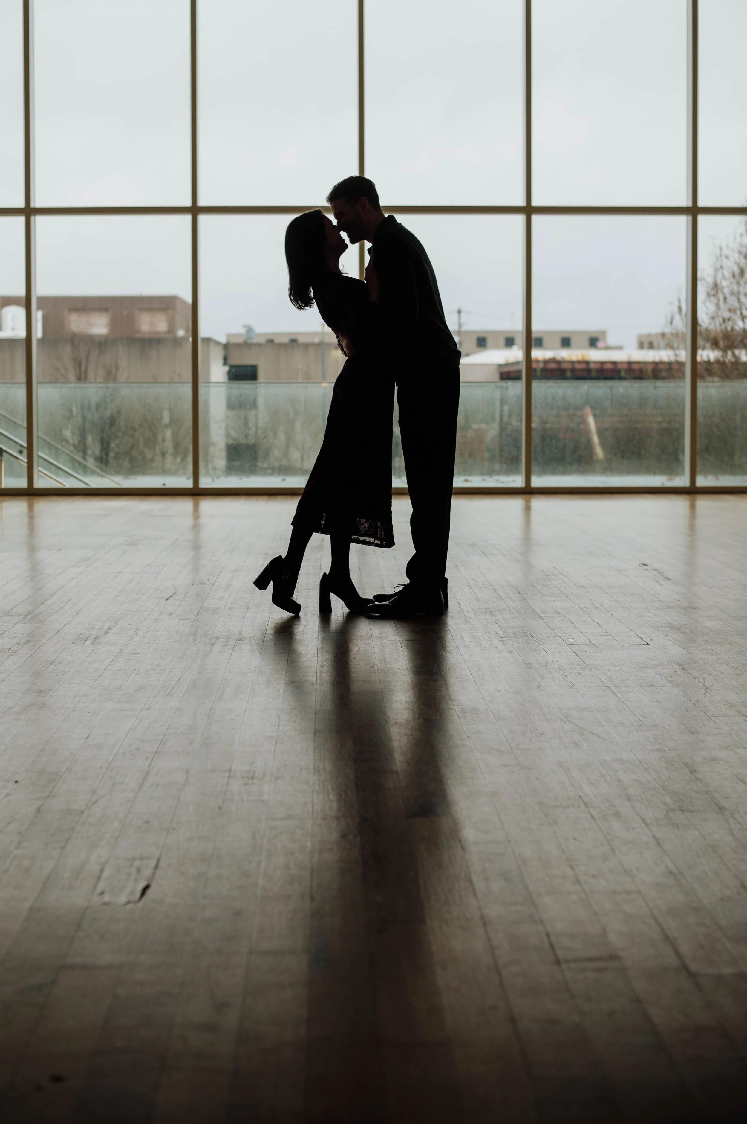 Silhouetted couple standing close in front of large windows, sharing an intimate moment indoors.