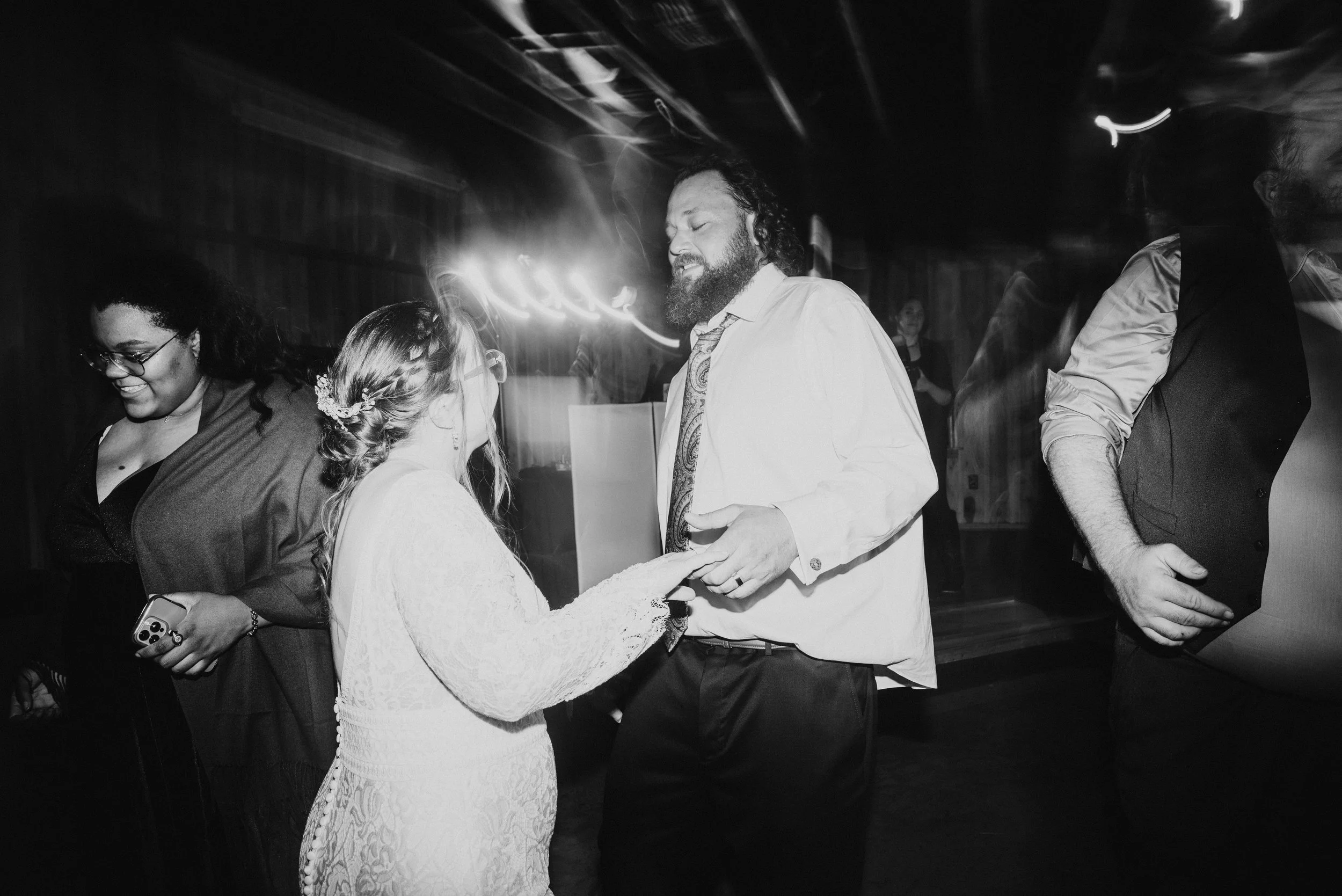 People dancing at a wedding reception, including a woman in a white dress and a man in a white shirt with a patterned tie, holding hands and dancing.