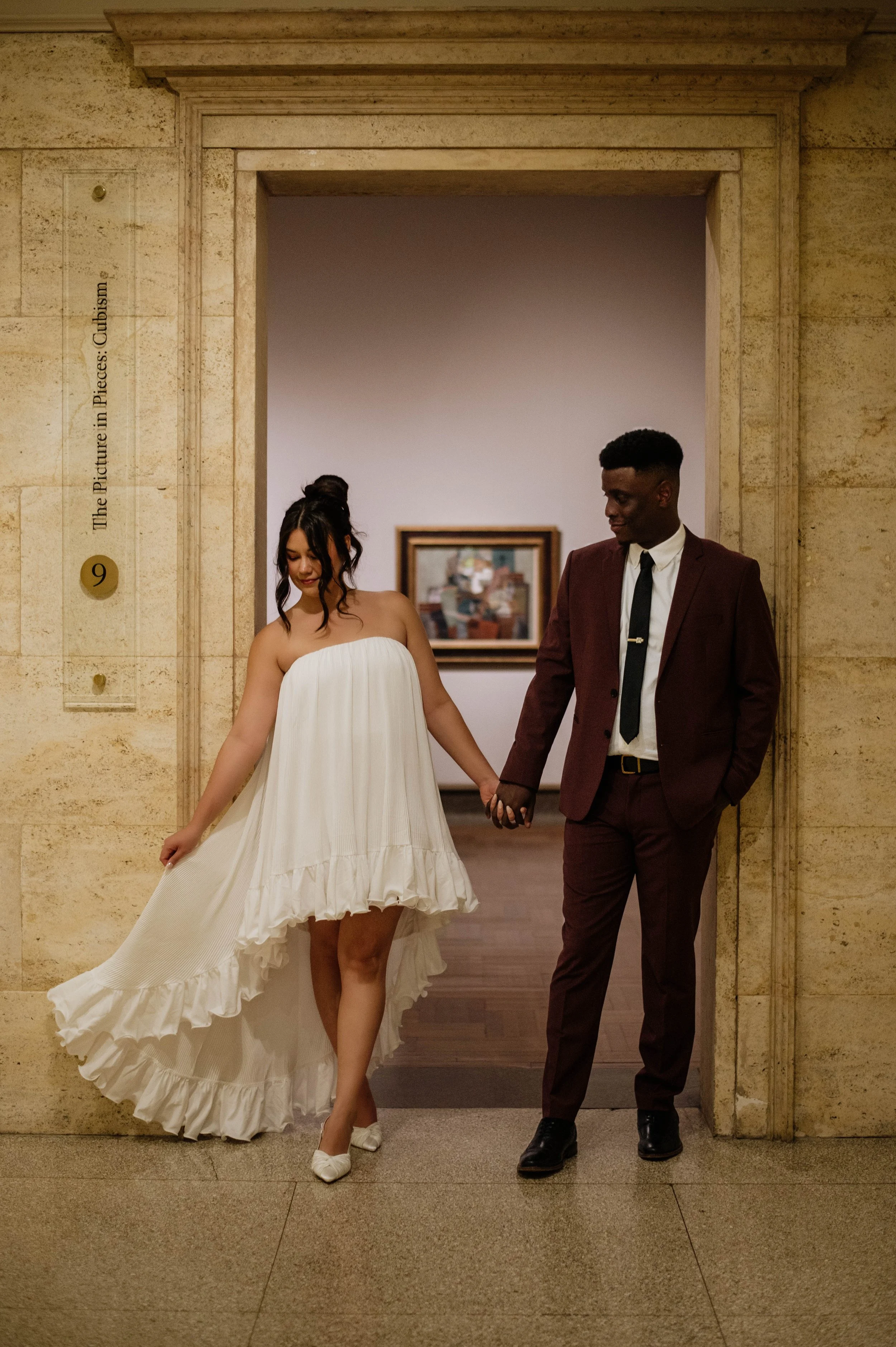A woman in a white strapless dress and a man in a maroon suit holding hands and walking through a doorway in an art museum.