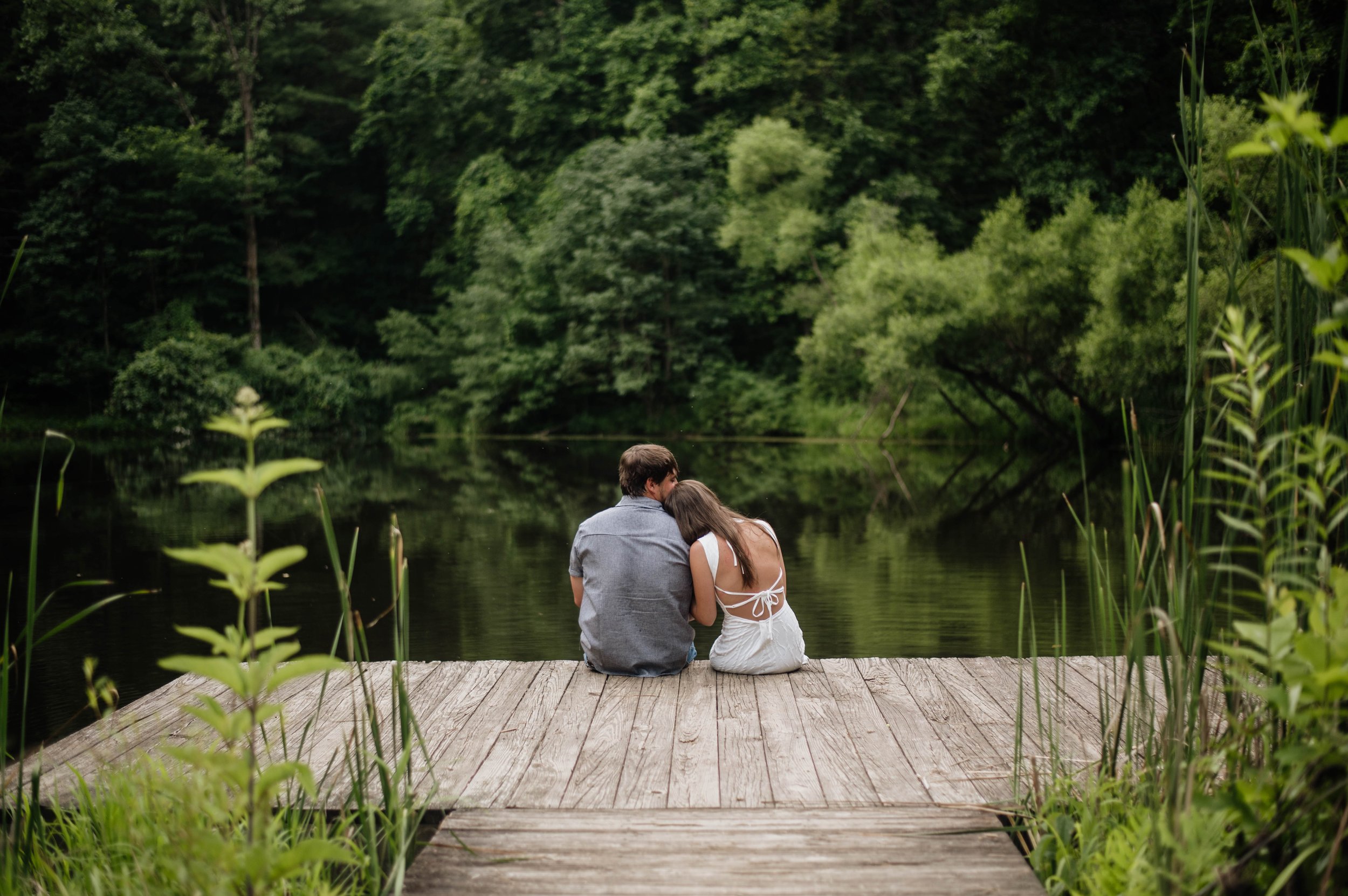 Engaged couple snuggle on a dock at a pond in the woods
