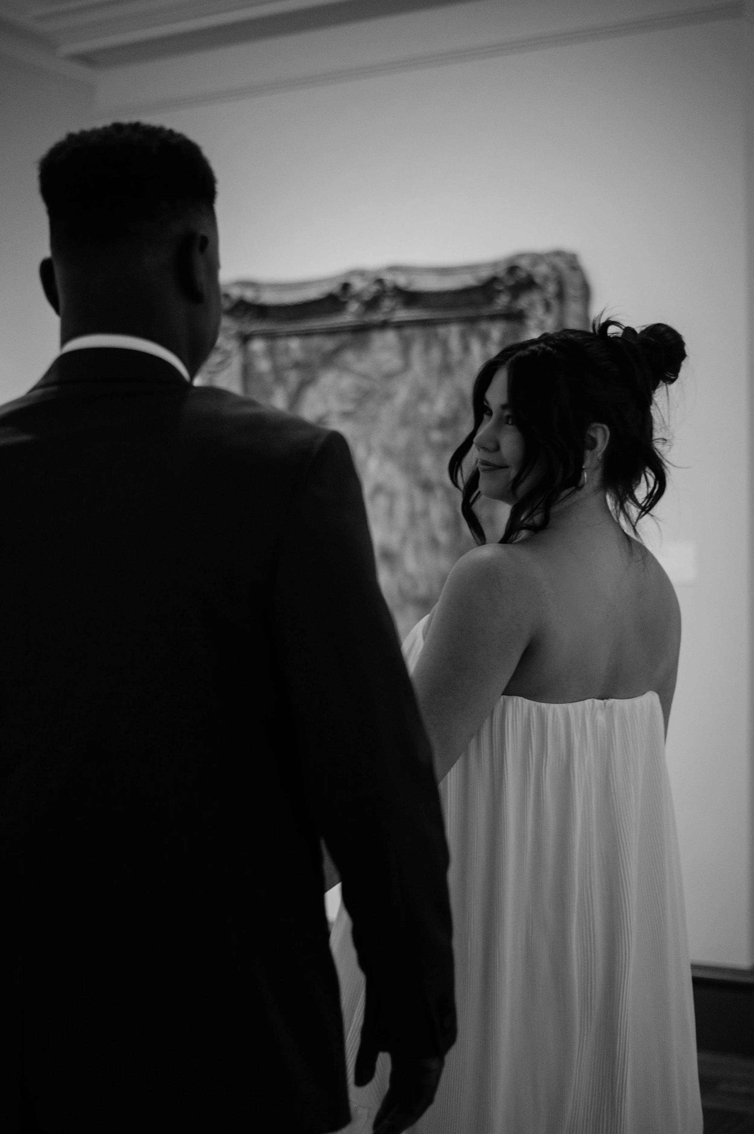 A black-and-white photo of a couple, with the man in a suit and the woman in a strapless dress, looking at each other; they are indoors with a decorative painting behind them.