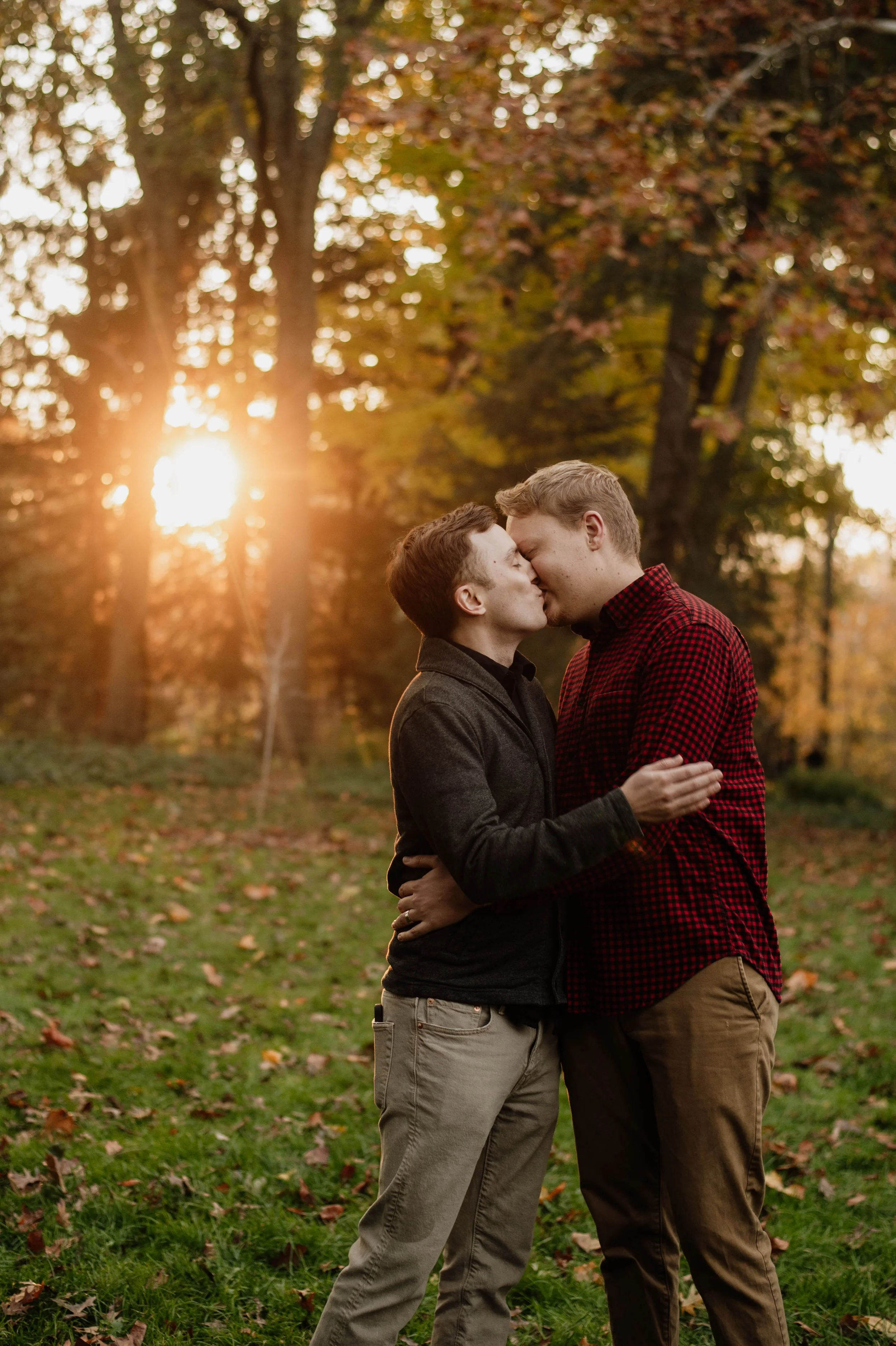 A couple kissing outdoors during sunset in autumn, surrounded by trees with fall foliage.