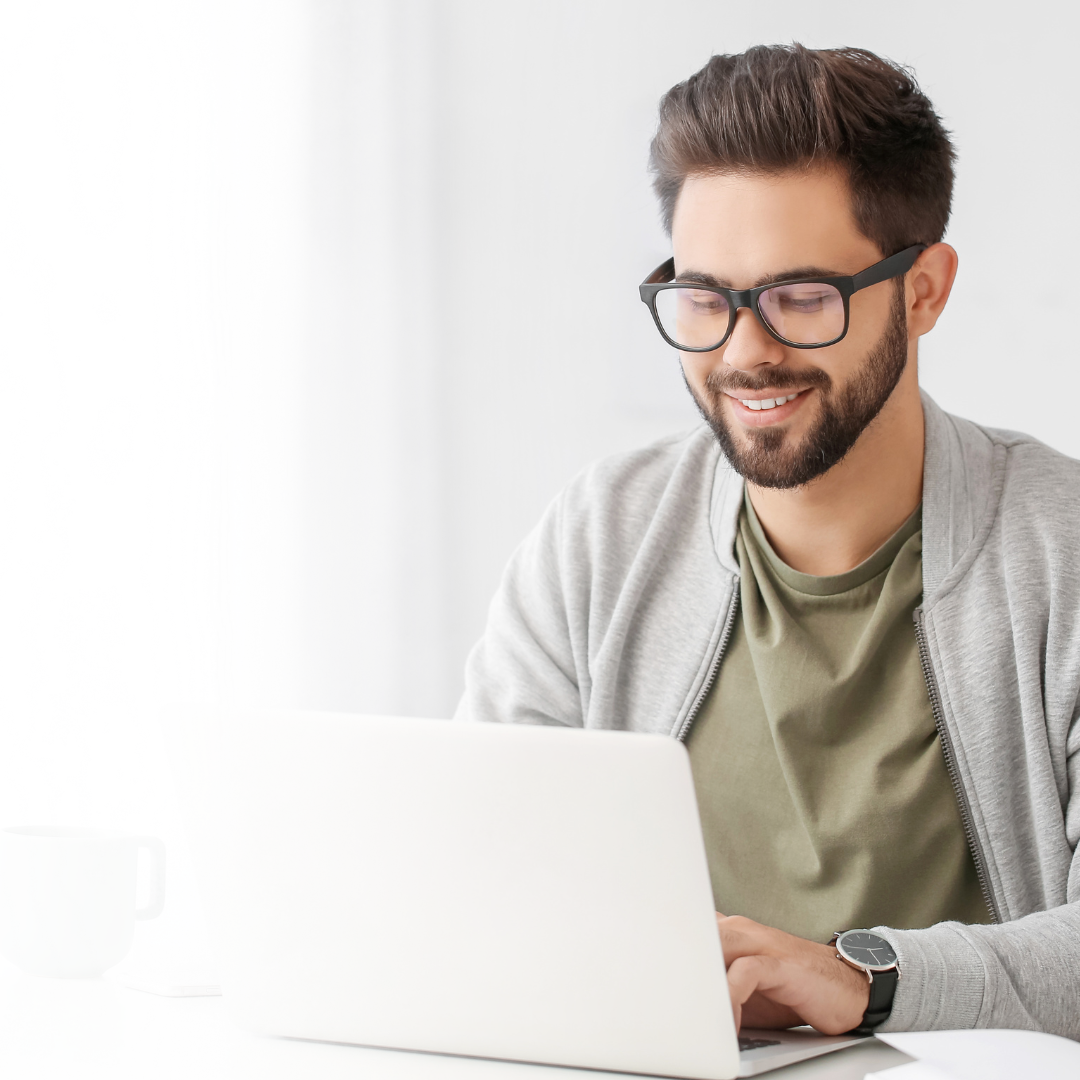 A man with glasses and a beard working on a laptop at a white desk, smiling and wearing a casual green T-shirt and gray jacket.
