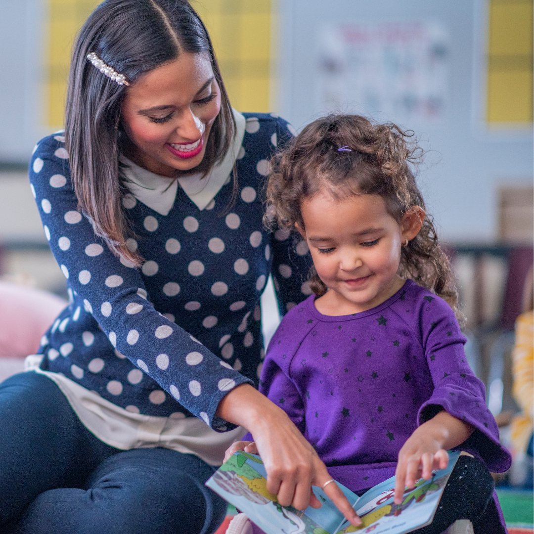 A woman with dark hair, wearing a navy blue polka dot sweater, is helping a young girl in a purple shirt read a colorful book in a classroom.