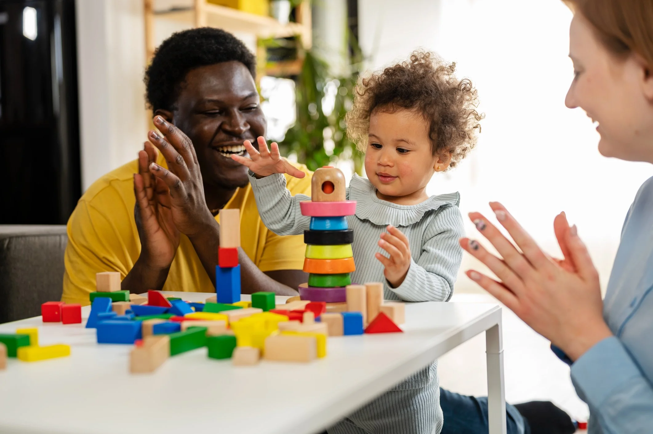 A smiling toddler stacking colorful wooden toys at a white table, with two women cheering and encouraging her.