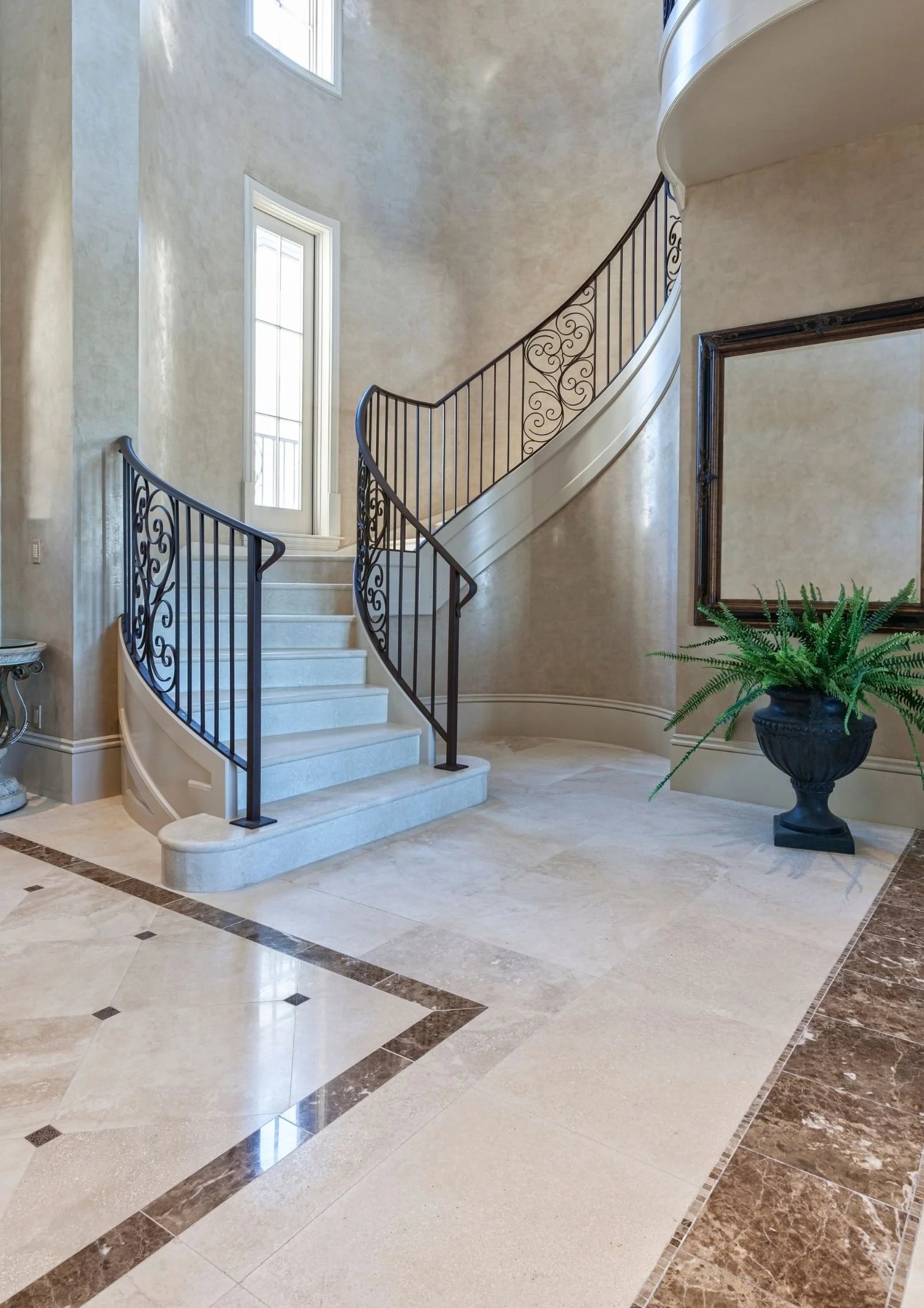 Elegant hotel or mansion interior with a curved staircase, wrought-iron railing, large window, potted fern, and decorative floor tiles.