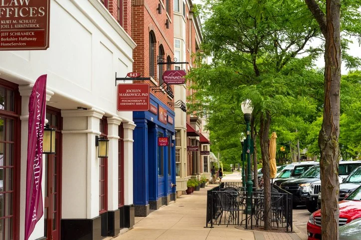 Street view with storefronts, trees, parked cars, and outdoor seating area with tables and chairs.