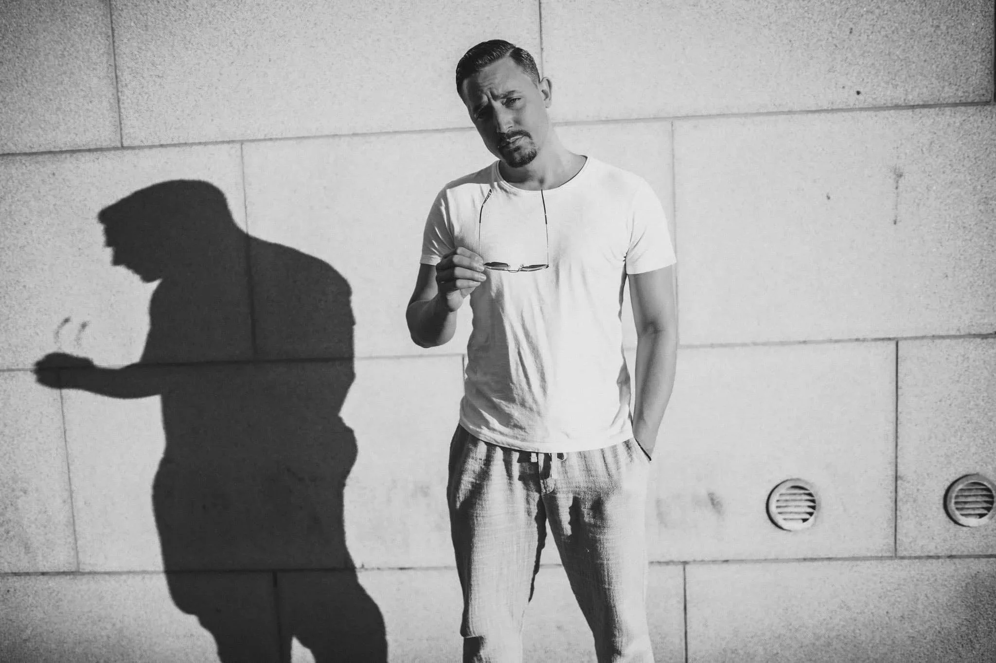 A man leaning against a wall in sunlight, holding glasses in one hand, with his shadow cast on the wall.
