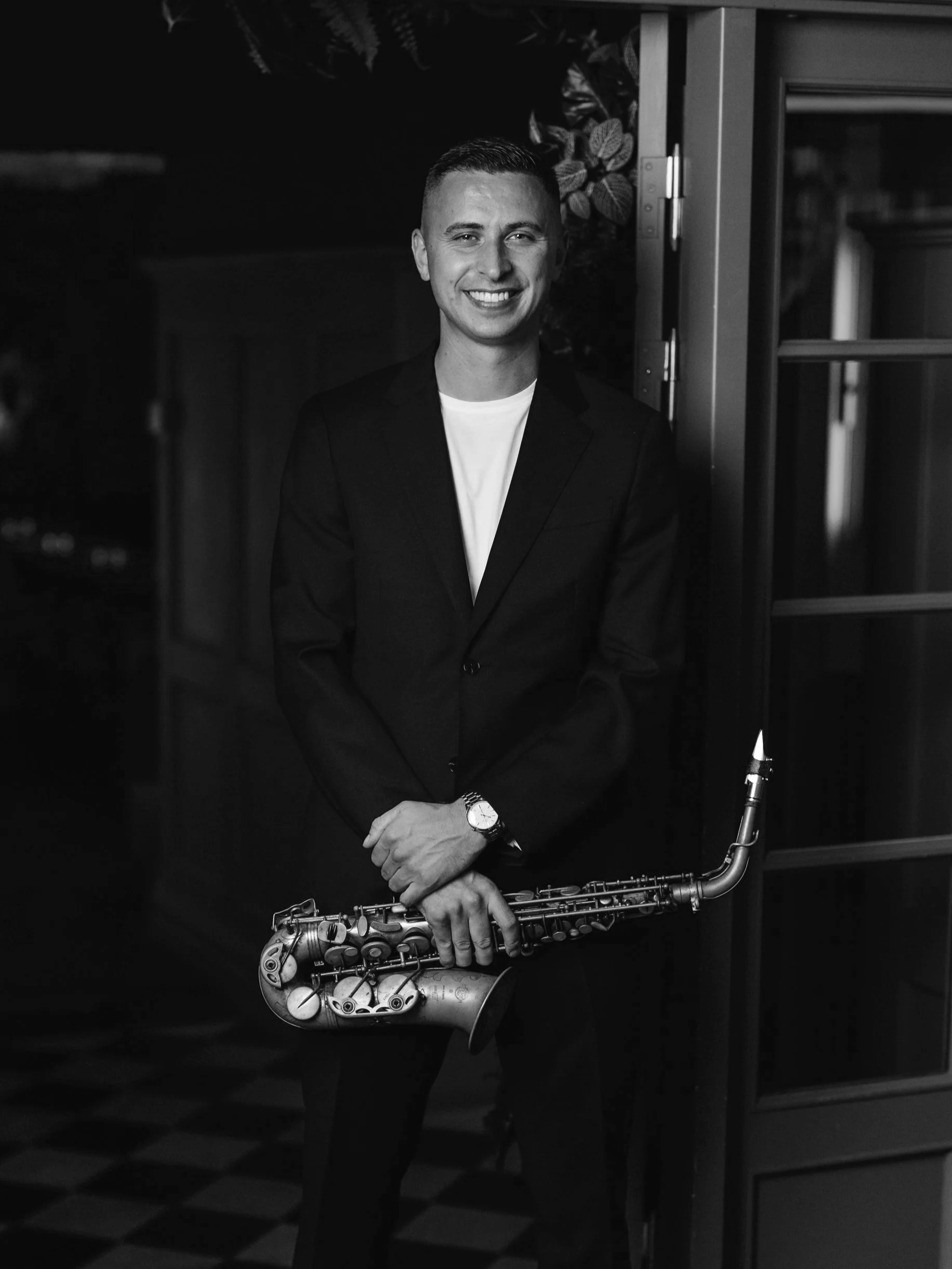 Black and white photo of a smiling man in a suit holding a saxophone, standing indoors near a glass door.