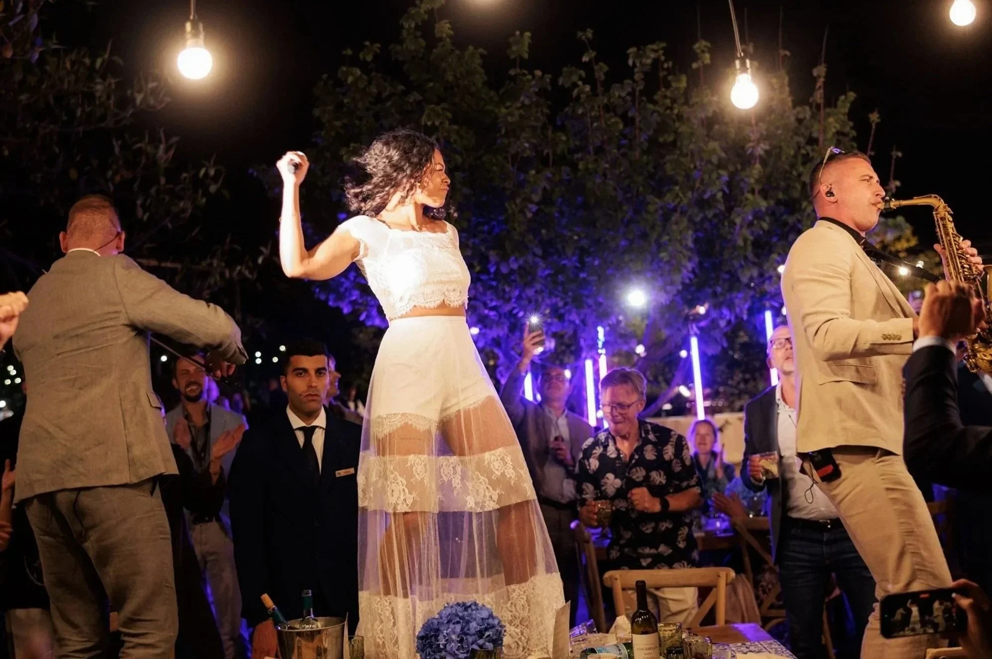 People at an outdoor party with string lights, dancing, and drinking, with a woman in a white lace dress dancing among friends.
