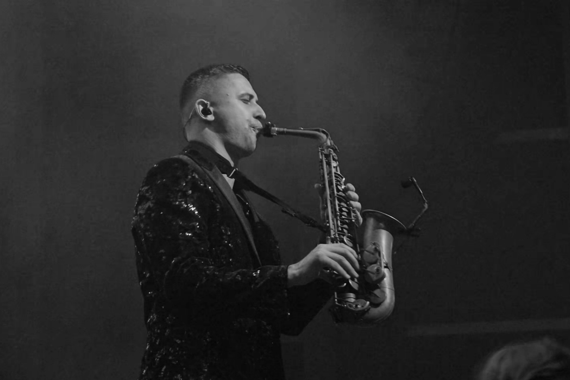 Man playing saxophone on stage, dressed in a shiny, sequined jacket, in black and white.