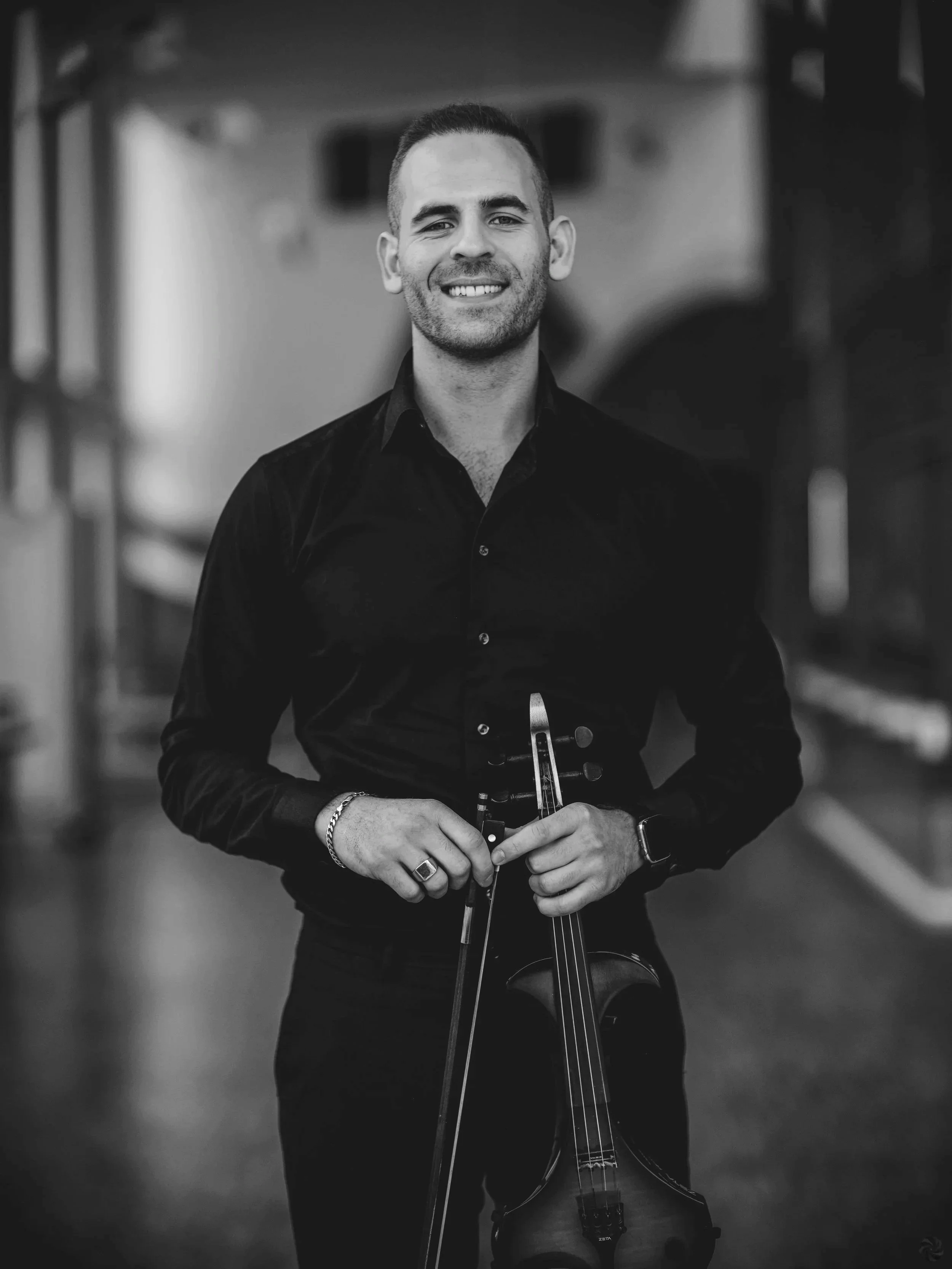 A man smiling and holding a violin and bow, standing indoors in a well-lit environment.