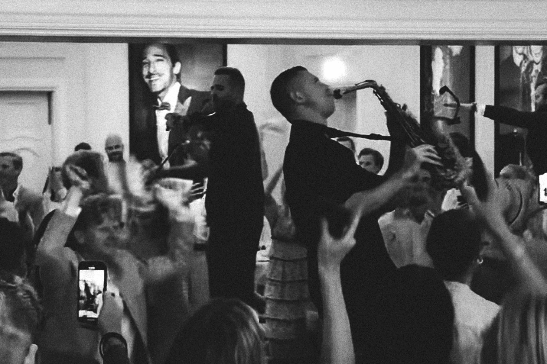 A black and white photo of a lively party with people dancing and enjoying live jazz music. Two musicians playing saxophone and violin are visible among the crowd.