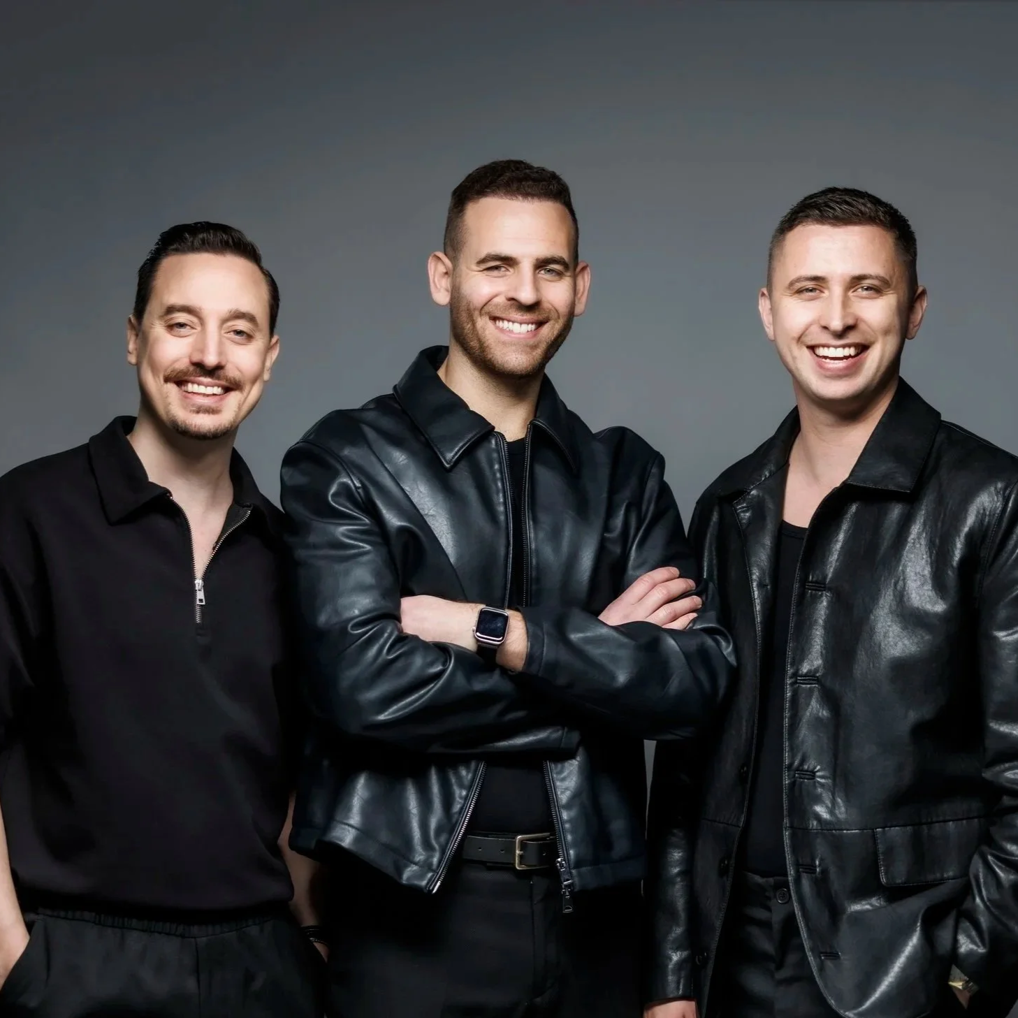 Three young men, SAX2VIOLIN, wearing black leather jackets and black shirts, standing together and smiling against a gray background.