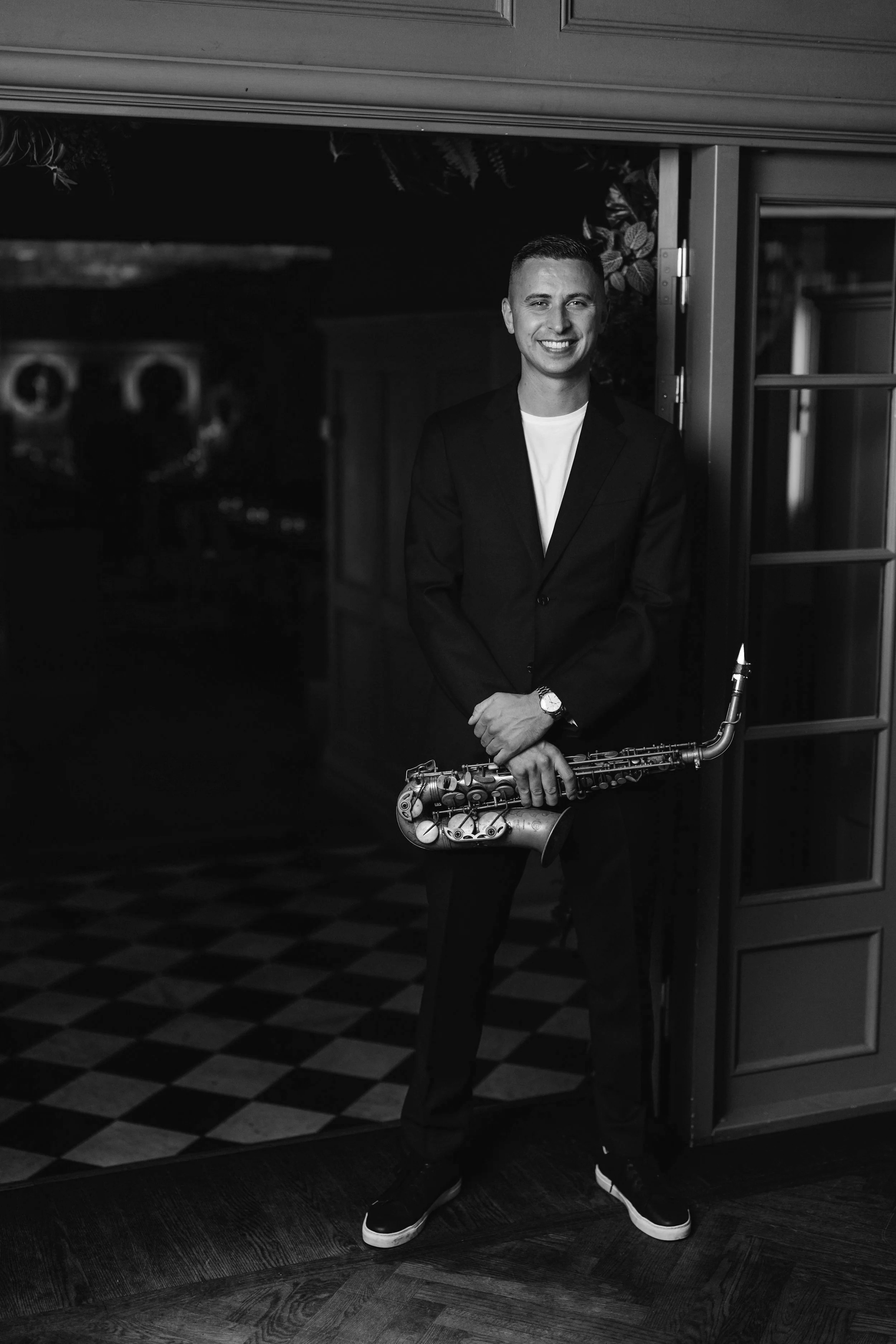 A man in a suit holding a saxophone, standing in front of a doorway in a dimly lit room with checkered flooring.