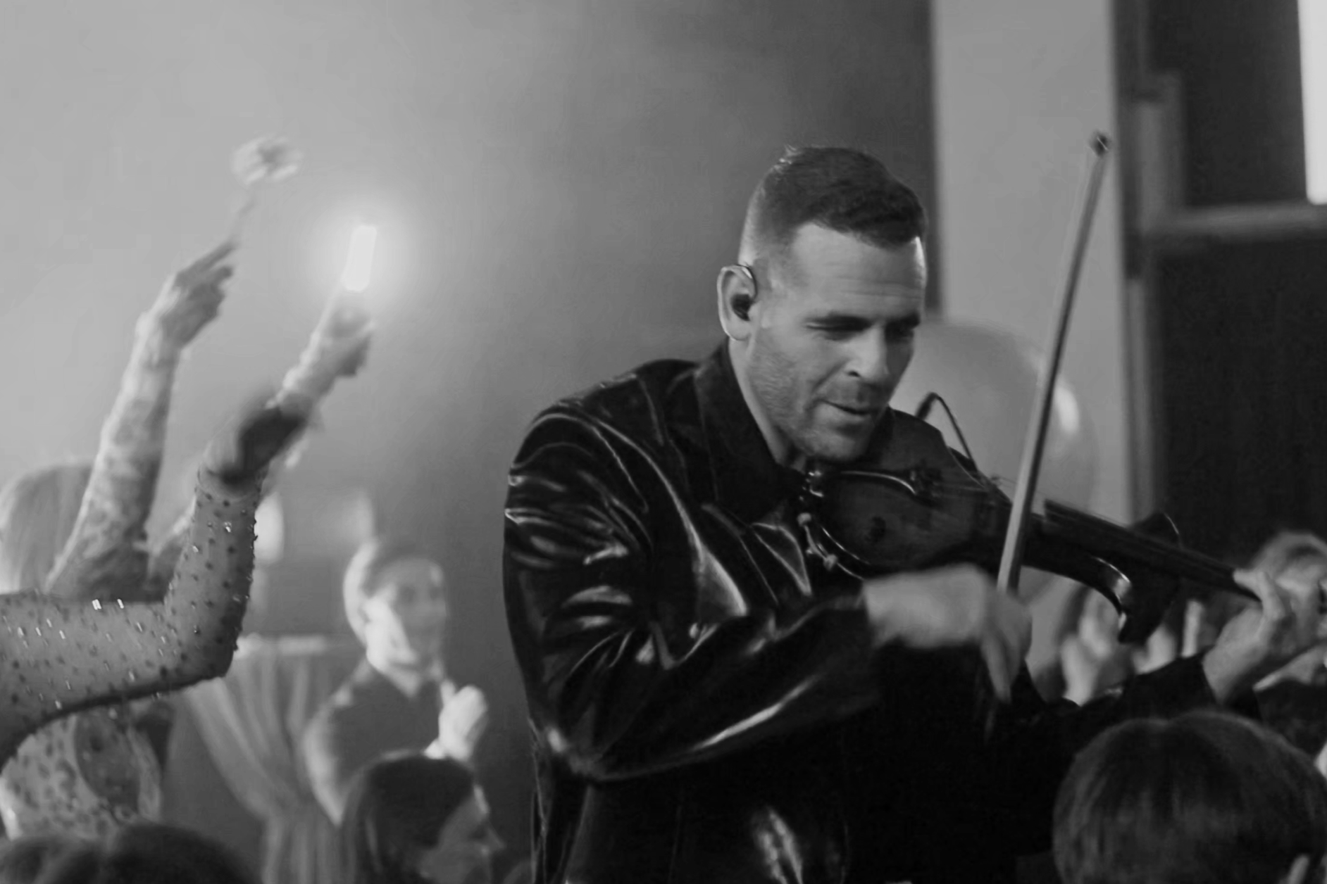 A man playing a violin in a crowded, lively setting, with spectators in the background and someone taking a photo or video with a smartphone in the air, black and white image.