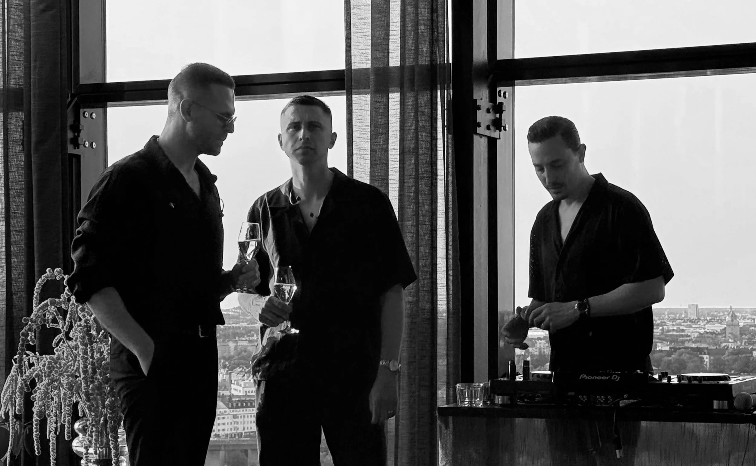 Three men in black shirts, two holding glasses of champagne, standing near a window with cityscape view, one using DJ equipment
