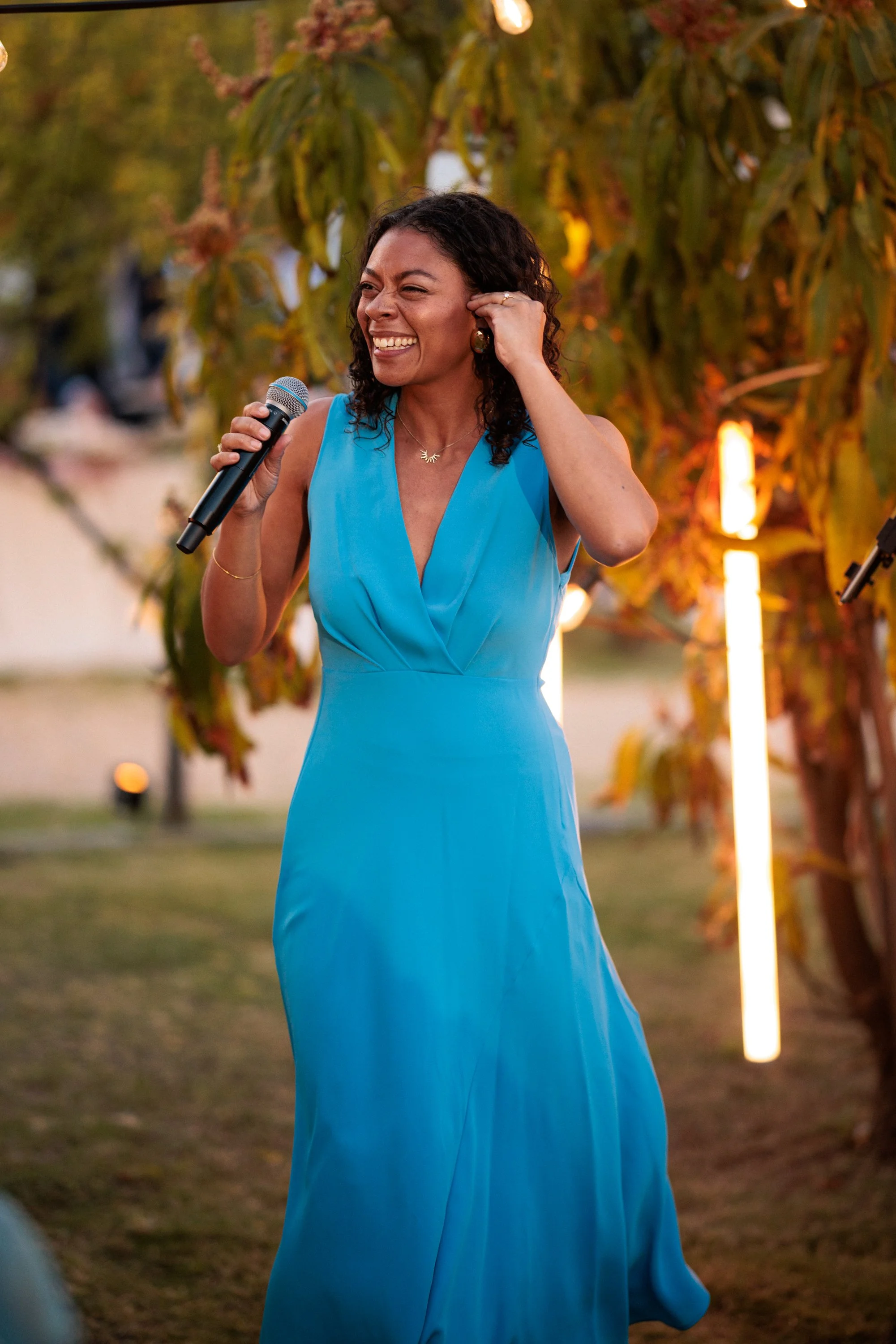 A woman in a blue dress smiling while holding a microphone, outdoors with trees and warm lighting in the background.
