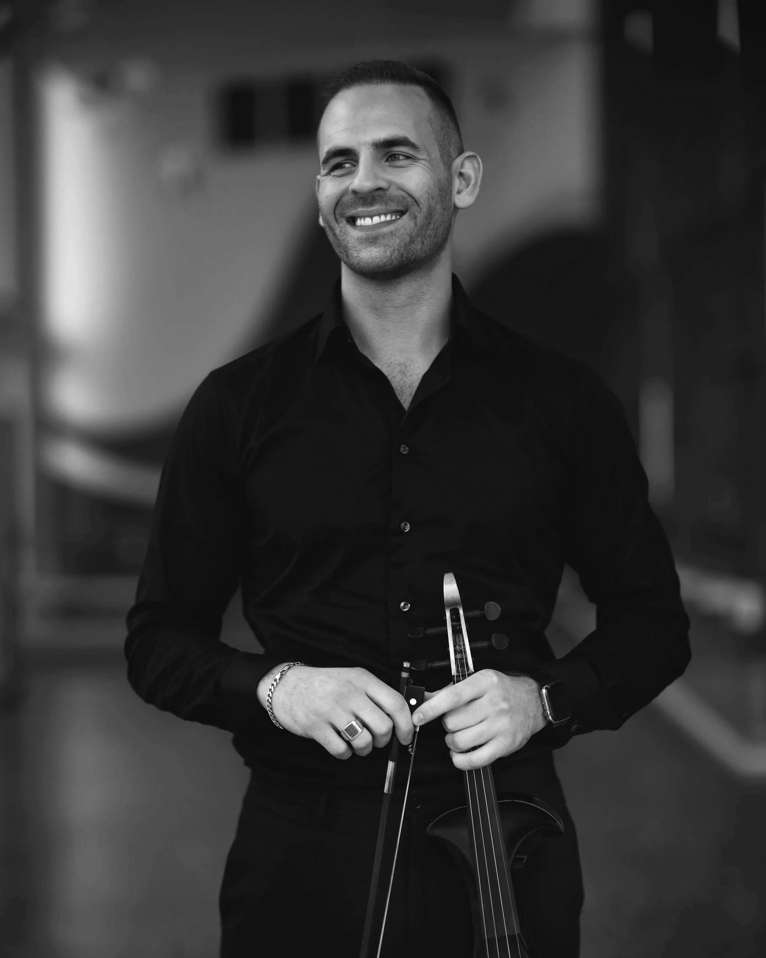 Black and white photo of a smiling man holding a violin, wearing a black shirt and accessories, in a blurred indoor setting.