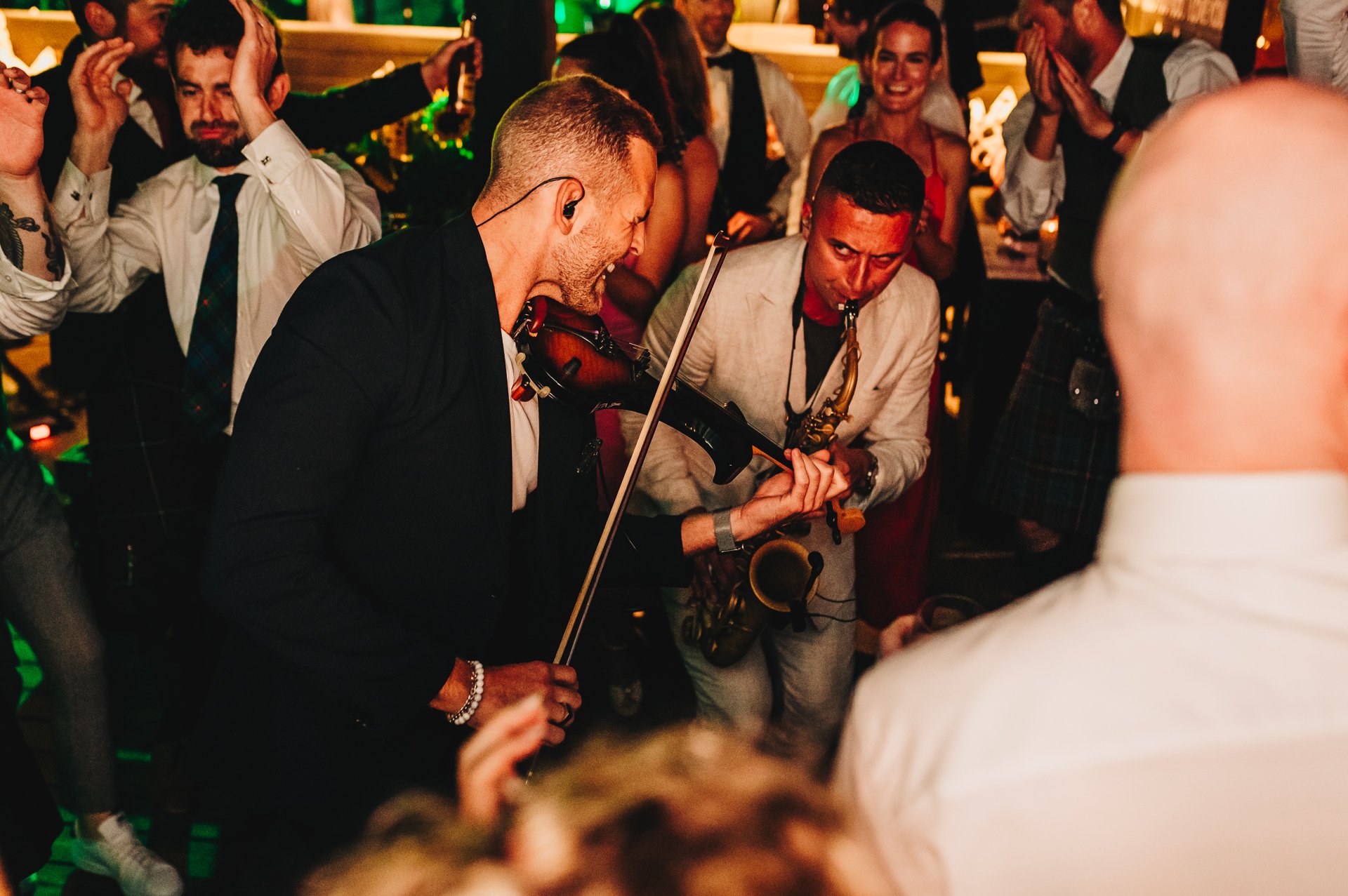 People dancing and enjoying live music at a party, with musicians playing violin and saxophone in the center.