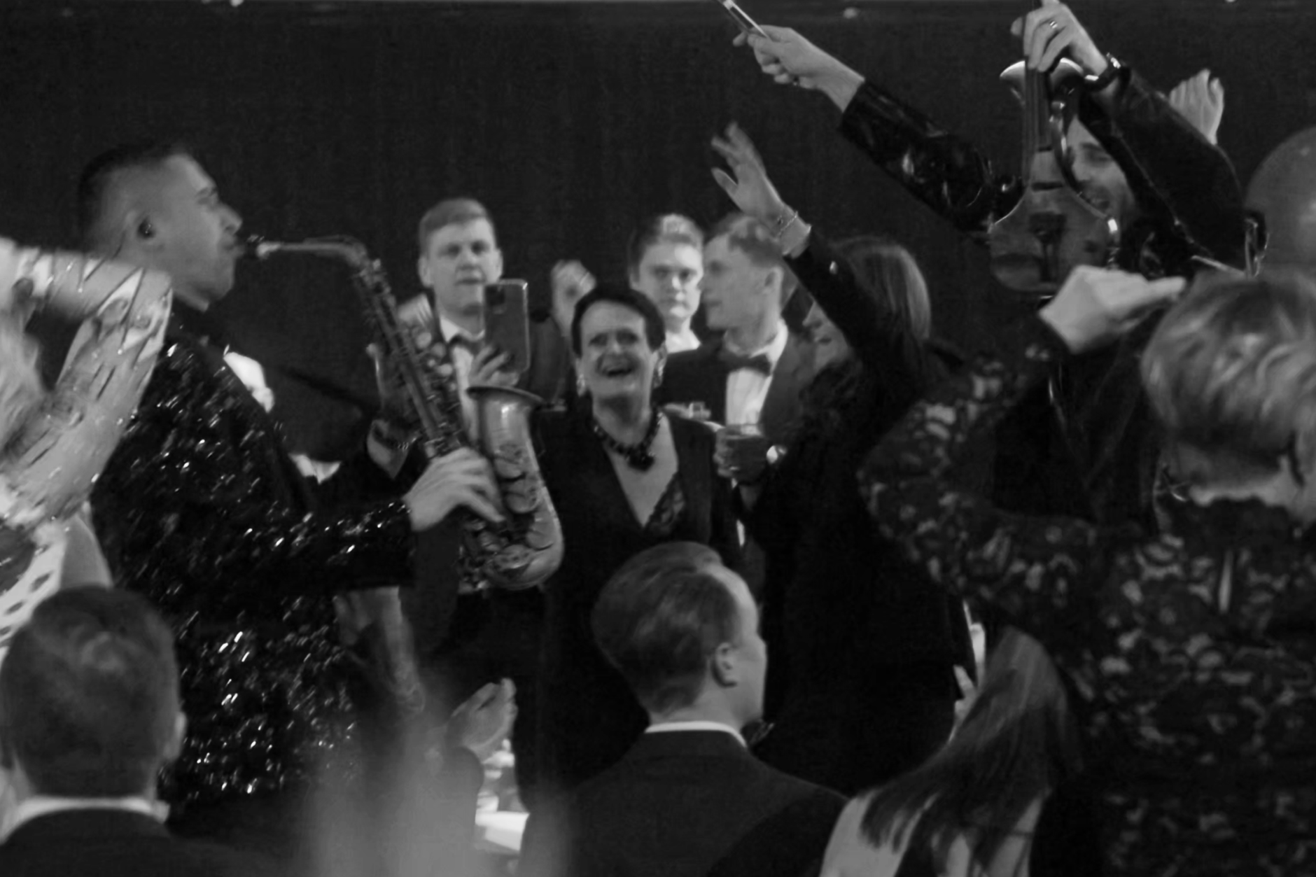 Group of people at a party, with some women dancing and smiling, and men in tuxedos, in a lively social gathering, with musical instruments and drinks.