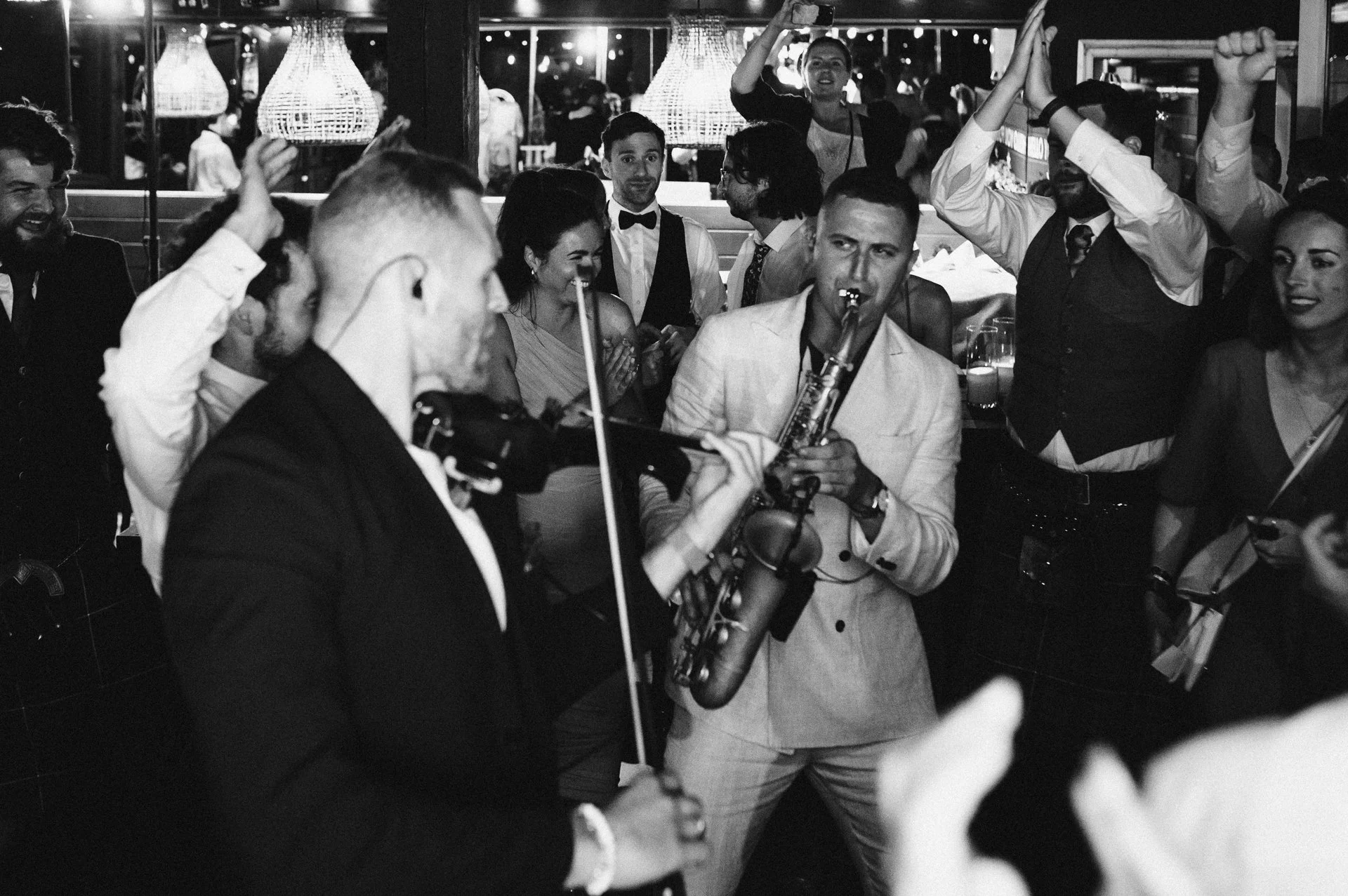 A lively black-and-white photo of a celebration with people dancing, playing musical instruments, and enjoying themselves in a festive setting.