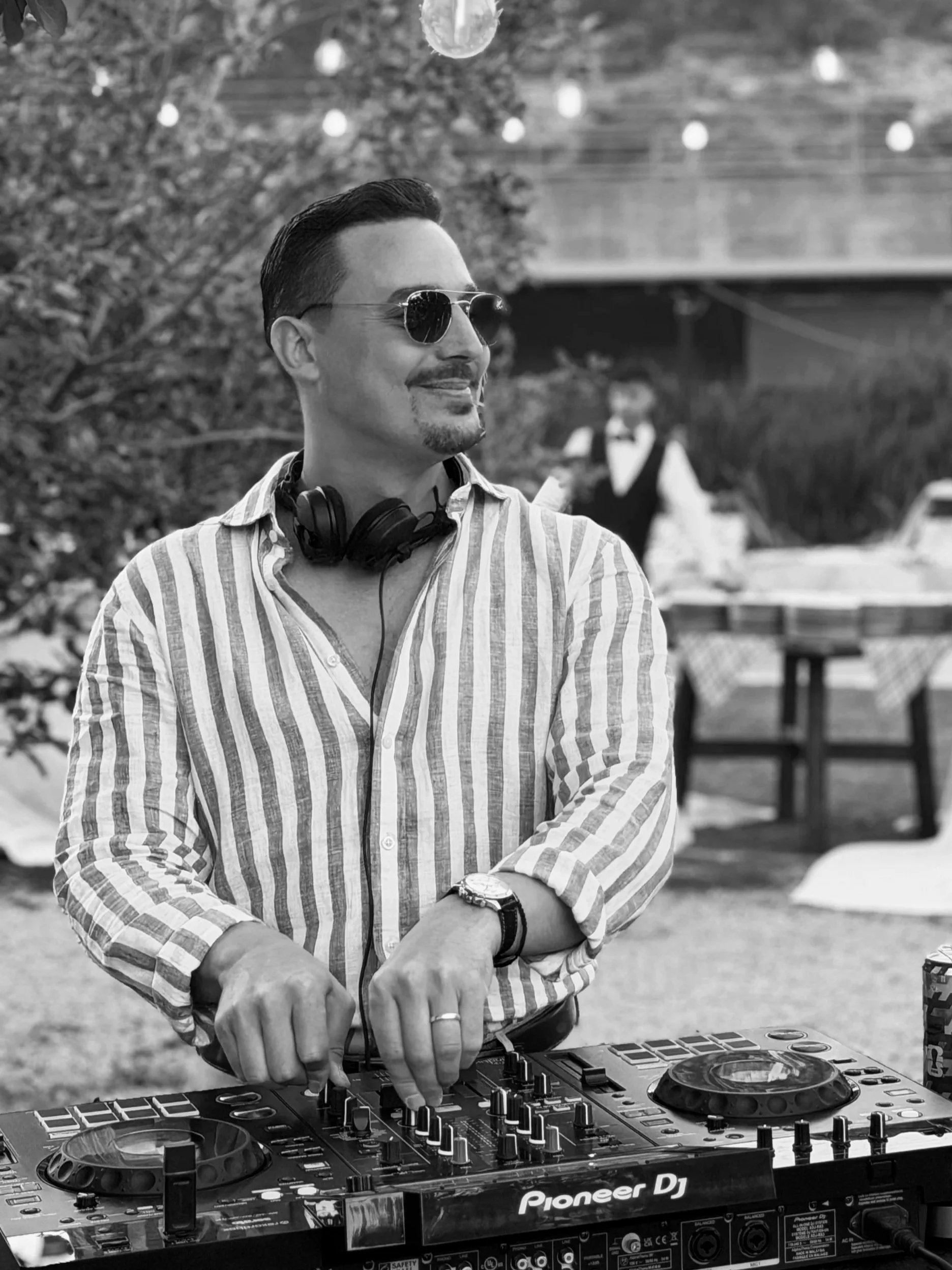 A man wearing sunglasses and a striped shirt is DJing with headphones around his neck, using a Pioneer DJ controller at an outdoor event.