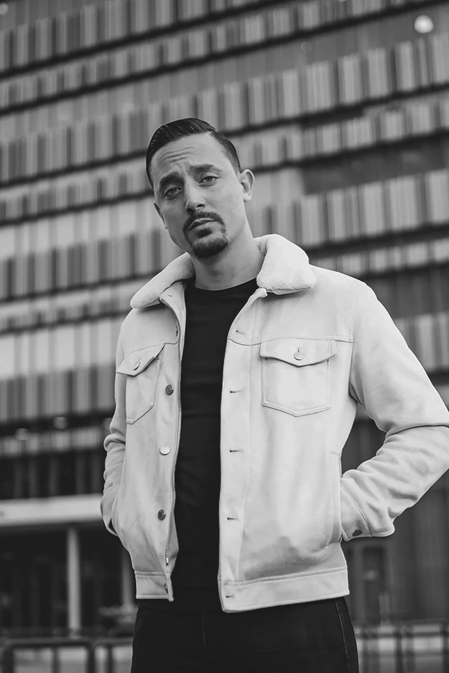 A man with a serious expression standing in front of a building with large windows, wearing a light-colored jacket over a black shirt.