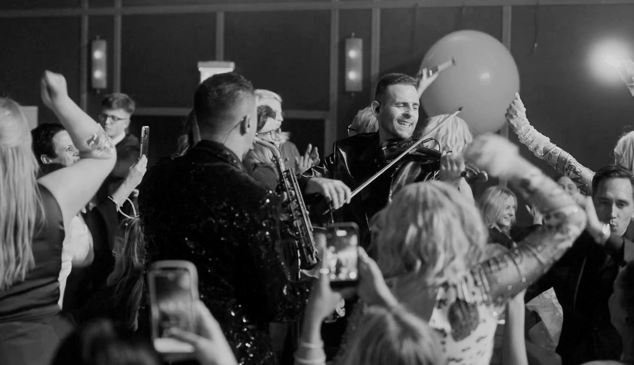 People dancing and enjoying live music at a celebration or party, with a violinist performing among the crowd, some capturing the moment on their smartphones.